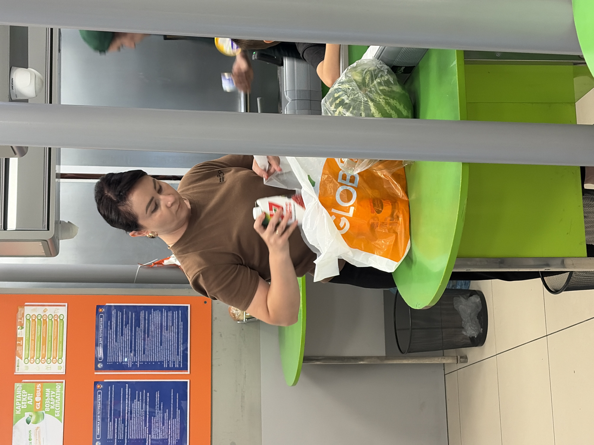 In the interior of what appears to be a grocery store, a woman with dark hair pulled back and wearing a brown t-shirt is standing at a green counter. She is holding a white carton with red accents in her right hand and reaching into a large orange plastic bag with her left hand, which contains other items. To her left, a black trash can is visible, containing crumpled grey plastic. In the background, another person wearing a green cap is visible, and a large watermelon wrapped in plastic is on the counter to the right of the woman. The scene is well-lit, suggesting it is daytime.