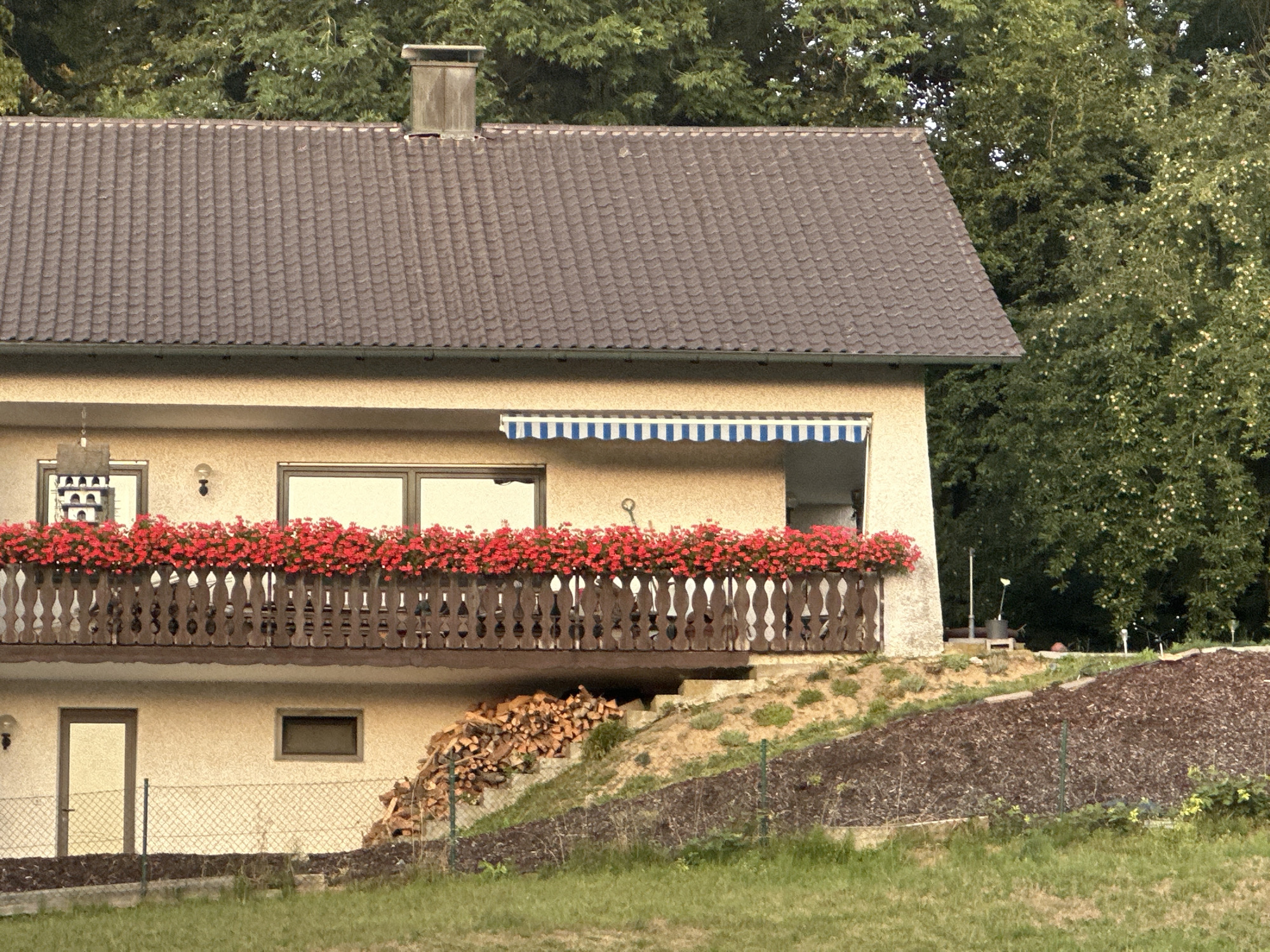 The image shows a close-up view of a two-story building with a brown tiled roof and a stucco facade. A wooden balcony with a railing filled with red flowers extends across the upper level. A blue and white striped awning is visible above a window on the upper story. A chimney is present on the left side of the roof. Below the balcony, there is a door, a small rectangular window, and a pile of firewood stacked against the wall. A chain-link fence runs along the bottom of the property, separating it from a grassy area and a sloped, mulched embankment. Lush green trees and foliage form the background. The scene is likely a residential property, potentially in a rural or suburban setting, given the presence of firewood and the natural surroundings.