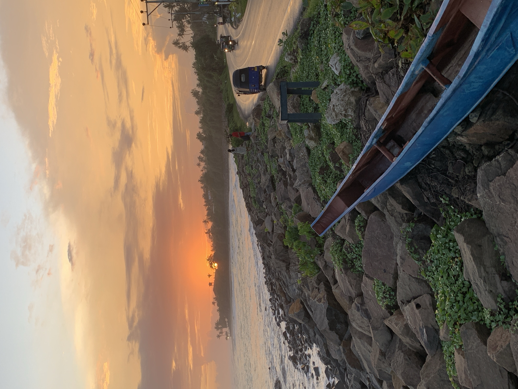 A view of the coast and sunset at Kathaluwa, Sri Lanka. The sun is setting over the water, a beach and palm trees are visible. A road with two vehicles runs along the coastline. In the foreground is a rocky embankment covered in vegetation with an overturned blue outrigger canoe or fishing boat resting atop. A small group of people stand along the beach by the water.