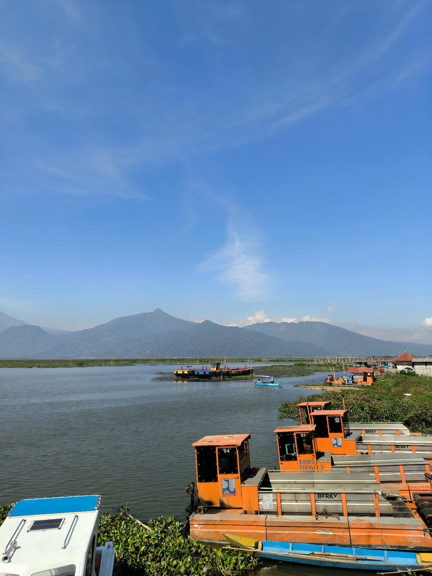 The image captures a serene waterfront scene in Tuntang, Indonesia, under a clear blue sky with wispy white clouds. In the foreground, several orange and white boats are docked along the shore, some laden with green aquatic plants. The orange boats, numbered "01," "04," and "12," have distinct cabins with dark windows. A white boat with a blue roof is also visible.
The middle ground features a wide expanse of calm water, reflecting the sky. Several other boats, including a small blue one with occupants, are scattered across the water. In the distance, a line of wooden poles stretches across the water, and beyond that, a verdant shoreline is dotted with small buildings and structures, one with a red roof.
The background is dominated by a range of majestic, hazy mountains, their slopes covered in vegetation. The overall impression is one of tranquility and natural beauty, with the boats suggesting human activity within a peaceful, natural environment.