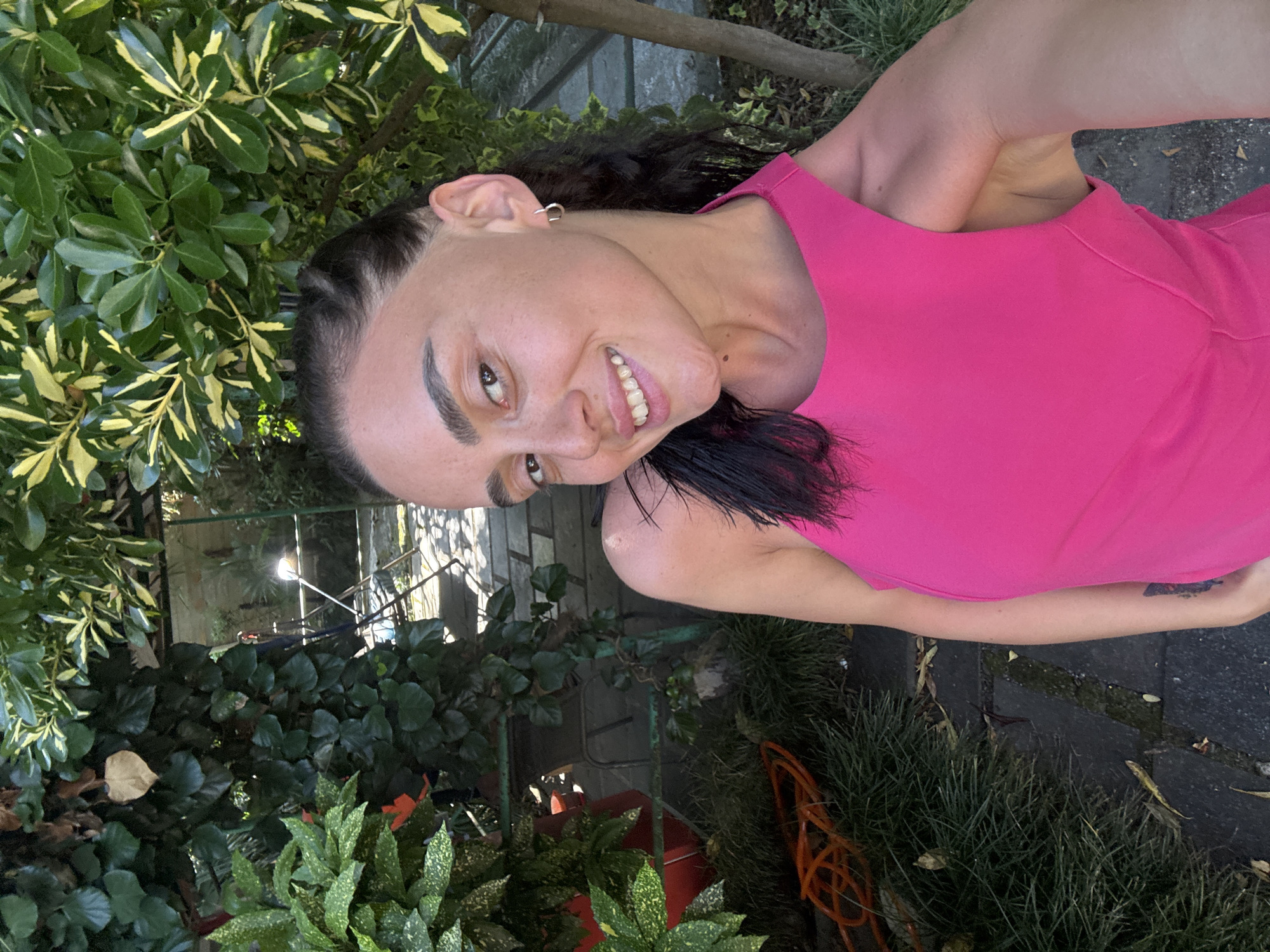 A young woman with dark hair pulled back into braids, is outdoors in a lush garden setting, possibly in Batumi, Georgia. She is wearing a bright pink, sleeveless top. Her head is tilted, and she is smiling widely, with her teeth visible. She has a silver hoop earring in her left ear.
The background is filled with dense green foliage, including leafy bushes and trees. Sunlight filters through the leaves, creating dappled light. There are also some visible elements of paving stones and a garden path, suggesting a residential or park-like environment. The overall impression is one of a warm, sunny day, with the subject appearing relaxed and happy.