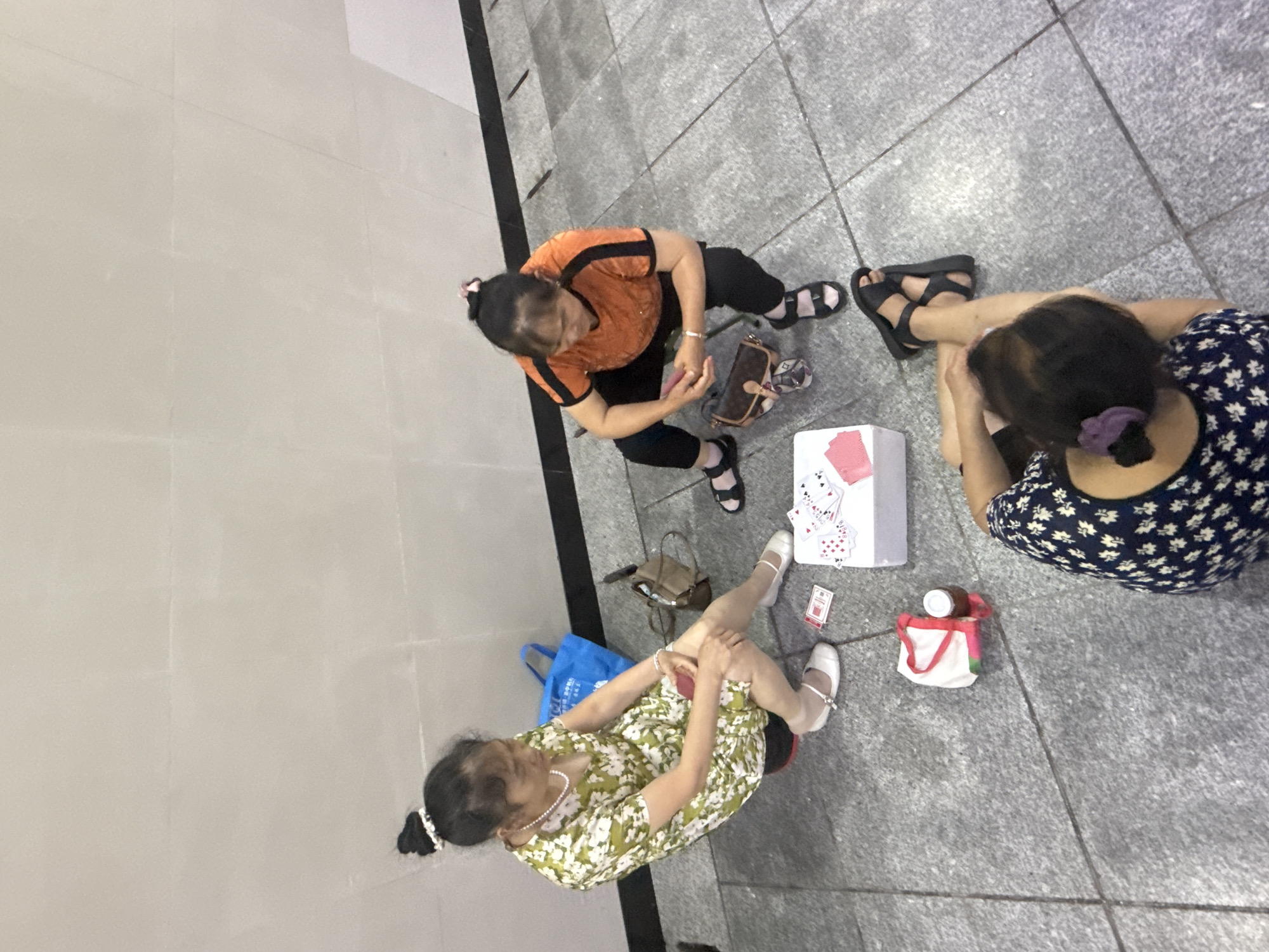 The image depicts three women sitting on a gray, tiled ground in an outdoor urban environment. The background is a light gray, textured wall with horizontal seams. The woman on the left is wearing a green and white floral dress and white Mary Jane shoes. She is holding a red object in her hands, and playing cards are scattered on the ground in front of her. Beside her is a blue tote bag. The woman in the middle is wearing an orange short-sleeved shirt with black stripes on the sleeves and black pants. She is also holding a red object and has a brown and beige handbag on the ground near her. The woman on the right is partially visible, wearing a dark blue and white floral top and black sandals. She is sitting with her back to the camera. In front of the woman on the left, there is a white box, likely containing playing cards, and a white bag with a red handle, and a bottle of liquid. The scene appears to be a casual gathering where the women are playing cards. The location is not explicitly stated to be Longta, China, but the setting is consistent with an urban public space in China.