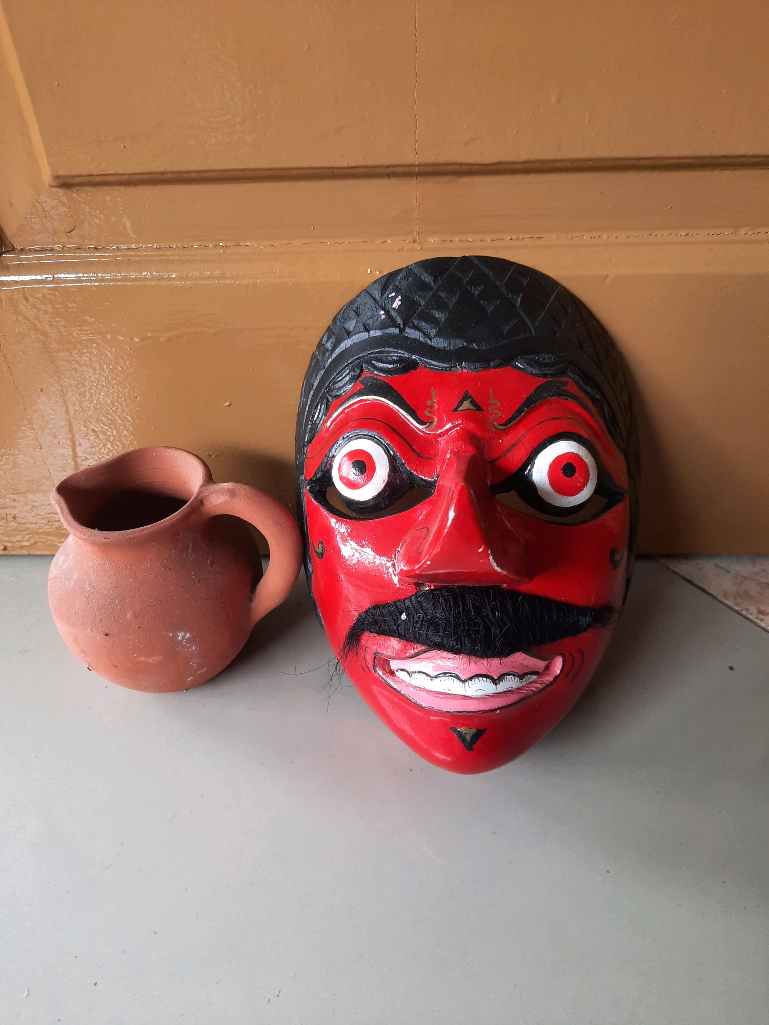 A red, theatrical mask with exaggerated features, including wide, staring eyes with red and white irises and black pupils, bushy black eyebrows, a prominent black mustache, and a wide, grinning mouth revealing white teeth, rests on a pale gray tiled floor. The mask has black hair painted on top and a black diamond pattern on the forehead. To the left of the mask is a small, unglazed terracotta jug with a handle. The background consists of a glossy, brown wooden surface with horizontal grooves, suggesting it might be a door or a cabinet. The lighting is direct, casting shadows. This scene is located indoors, possibly in a home in East Jakarta, Indonesia.