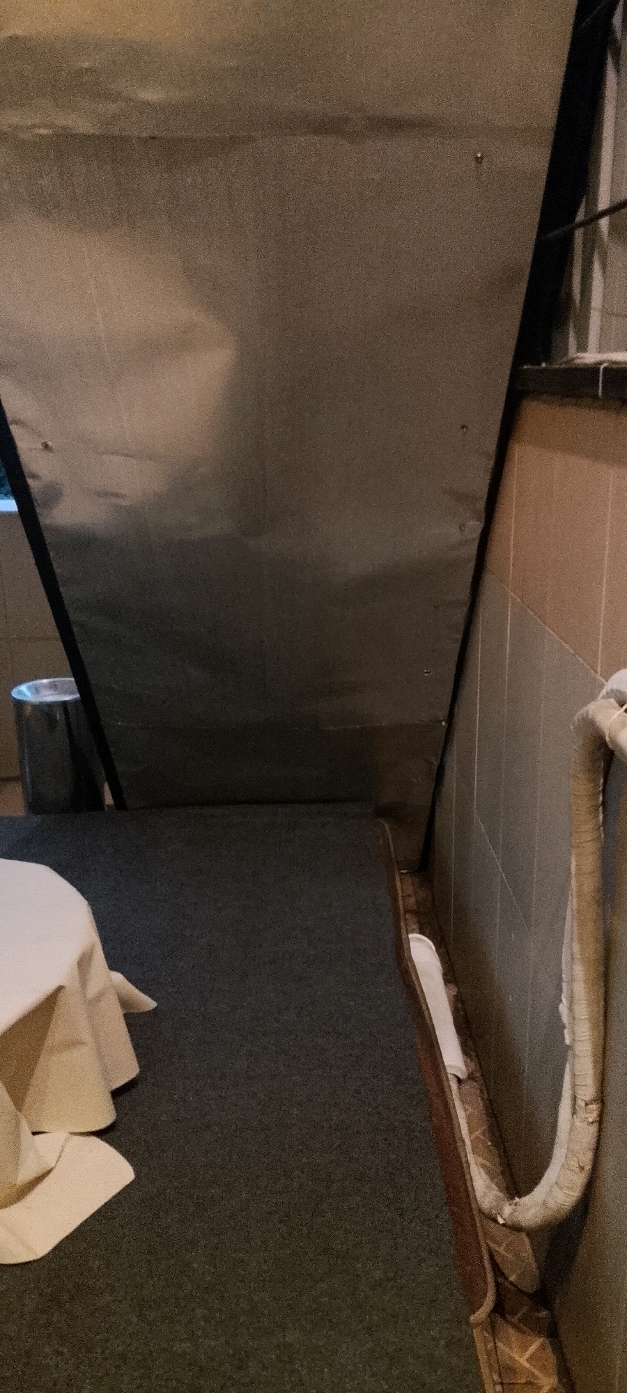 The image shows a dimly lit, indoor scene, likely a back room or storage area. In the foreground, a portion of a white tablecloth draped over a round object is visible on the left. The floor is covered in dark gray carpet. A large, metallic gray partition or screen dominates the upper half of the image, angled downwards. To the left of the partition, a silver trash can with a black rim is partially visible. The background wall is tiled with rectangular, light brown and gray tiles. A white pipe or conduit runs along the wall on the right side of the image. The overall impression is utilitarian and somewhat cluttered. There are no people or animals present. This scene does not provide any specific visual cues to identify its location within the city of Medan, Indonesia.