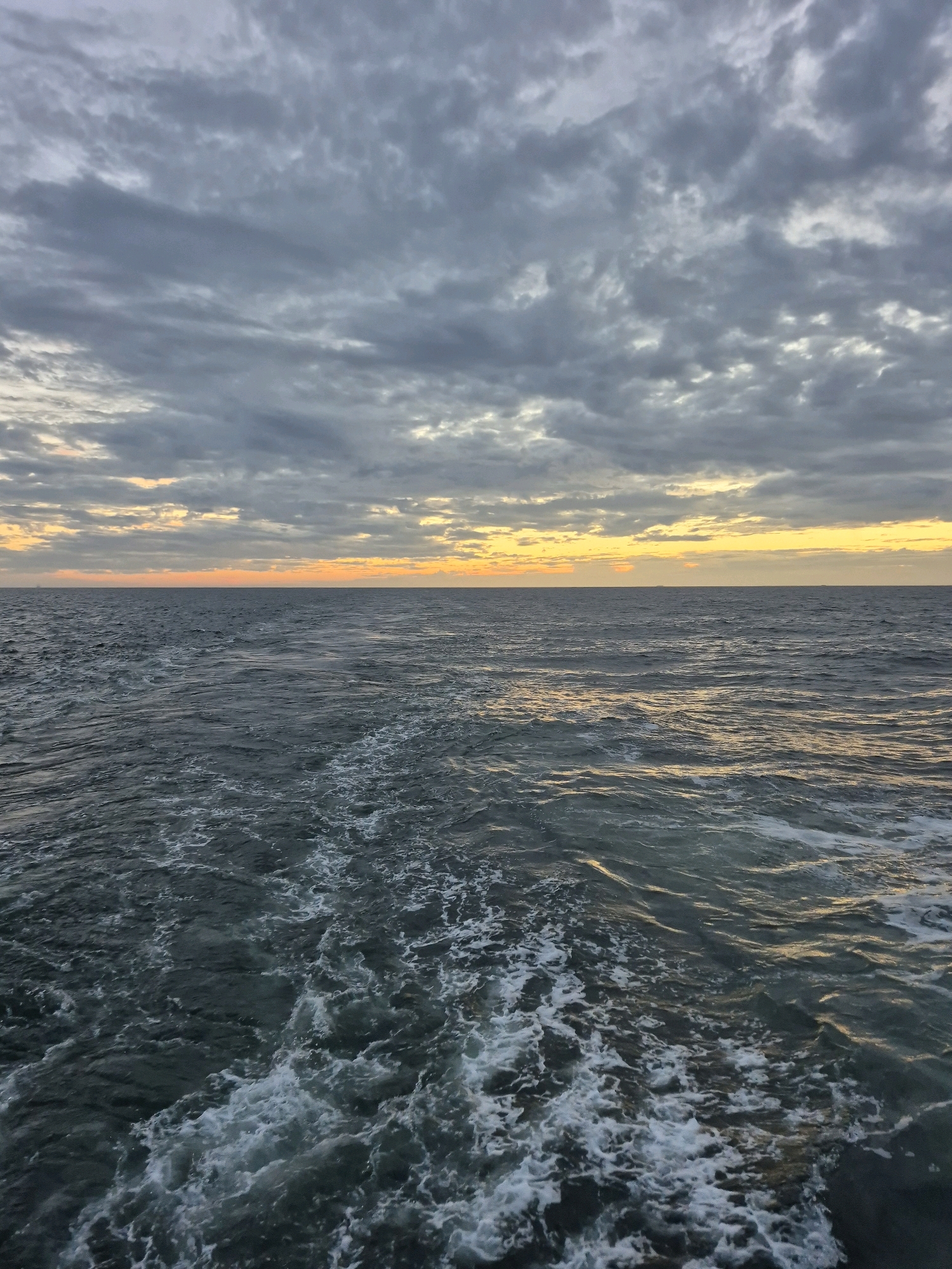 The image depicts a vast expanse of dark, choppy ocean water, disturbed by the wake of a vessel. White foam and spray are visible churning in the foreground, trailing away into the deeper water. The horizon is clearly defined, separating the sea from the sky. The sky is filled with heavy, grey clouds, but a band of bright, warm yellow and orange light is breaking through along the horizon, suggesting either sunrise or sunset. No people or specific structures are discernible. The scene appears to be at sea, far from any identifiable land features or urban environments within the city of Unknown, Germany, or elsewhere.