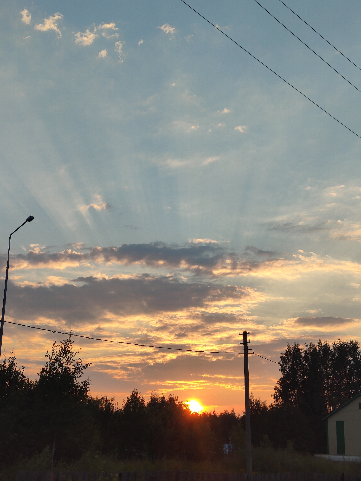 A low-angle, vertical shot captures a sunset over a treeline in Vysoky, Russia. The sky is a gradient of blue and orange, with rays of light emanating from the setting sun, which is partially obscured by the trees. Scattered clouds, tinged with orange and gray, dot the sky. In the foreground, a dark treeline forms a silhouette against the bright sky. To the left, a tall, dark lamppost stands at an angle. To the right, a power pole with wires stretches across the frame. A portion of a light-colored building with a green door is visible on the far right. A dark fence runs along the bottom of the image.