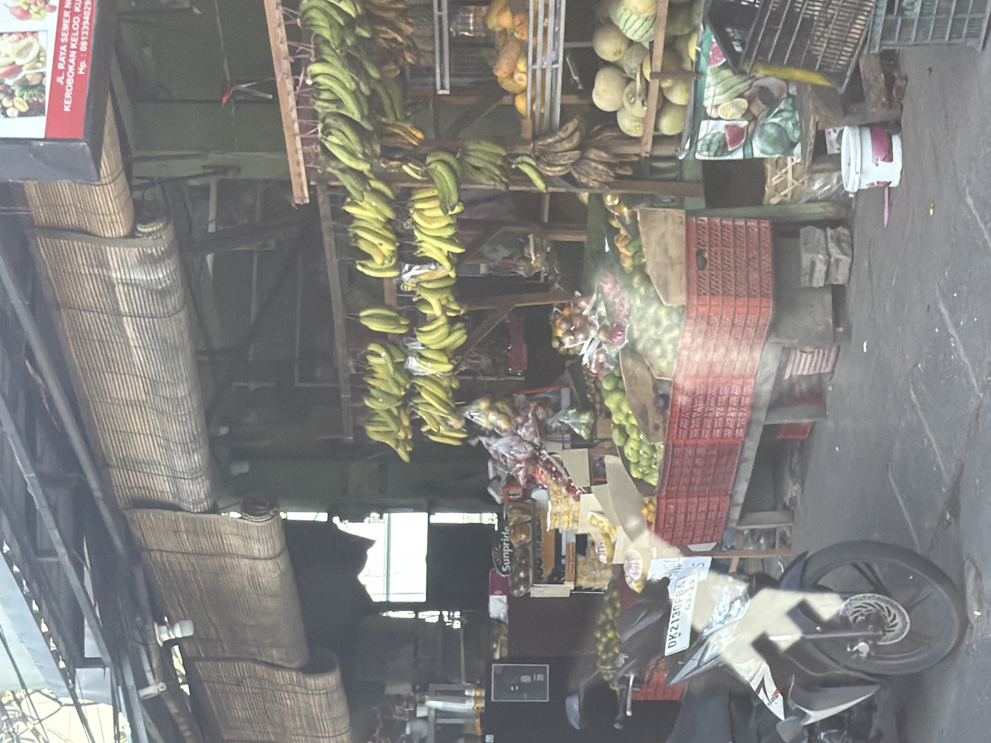 The image shows a fruit stand located on Jl. Raya Semer in Kerobokan Kelod, North Kuta, Indonesia. Bunches of green bananas are hanging from the roof of the stand. Additional fruits are arranged on the shelves and in crates. A motorcycle is parked in front of the stand. A sign displays the address and phone number.