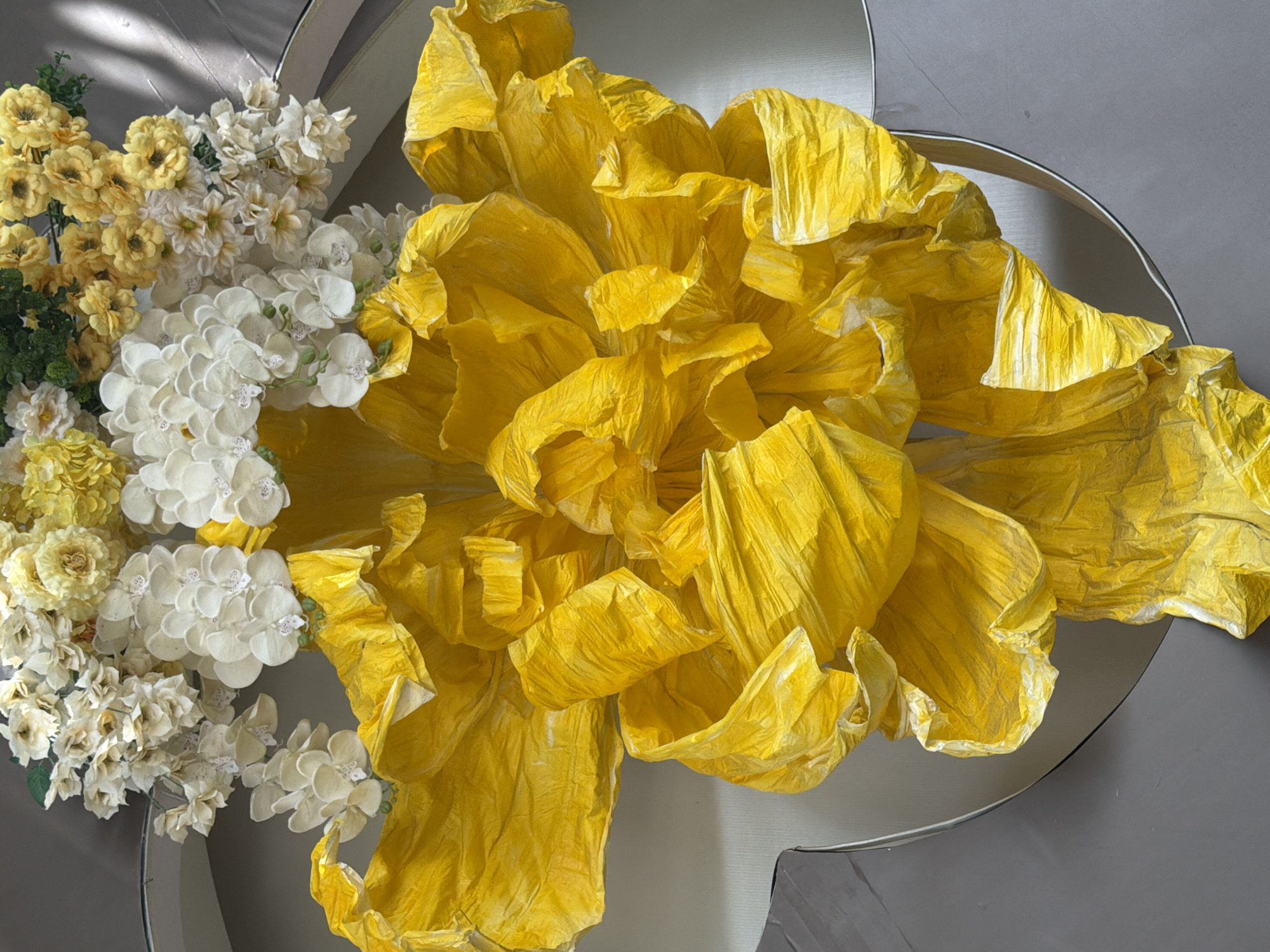 An overhead, close-up shot captures a vibrant, oversized artificial yellow flower crafted from crinkled paper, its petals dominating the foreground. The flower appears to be a centerpiece or decoration, possibly within a venue or public space. Behind the large yellow bloom, a cluster of smaller, artificial white and yellow flowers, including delicate white orchids, are arranged. The backdrop consists of a neutral gray surface with subtle shadows, suggesting an indoor setting or a shaded outdoor area. The overall impression is one of artistic display and floral arrangement. The image does not provide any specific visual cues to indicate its location in Nha Trang, Vietnam.
