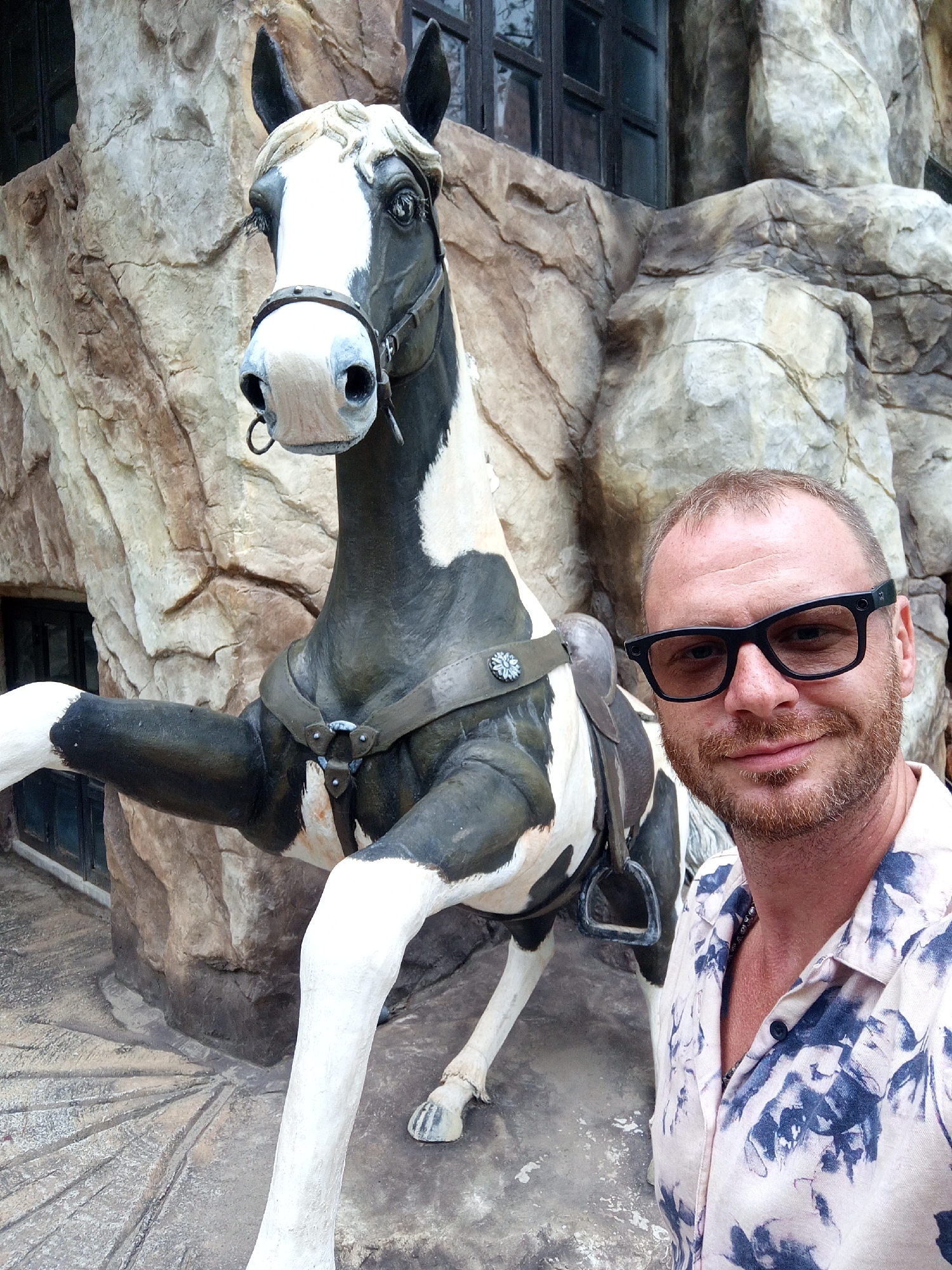 A man wearing glasses and a patterned shirt stands next to a statue of a black and white horse. The horse statue is in front of a large, rocky structure that resembles a cave or cliff face, with a dark-paned window visible. The man is smiling and looking at the camera. The ground in front of the statue is patterned with radiating lines. The scene appears to be outdoors, possibly in a themed park or tourist attraction. This appears to be the entrance to the Datanla Waterfall in Da Lat, Vietnam, which features a roller coaster ride with horse-themed cars.