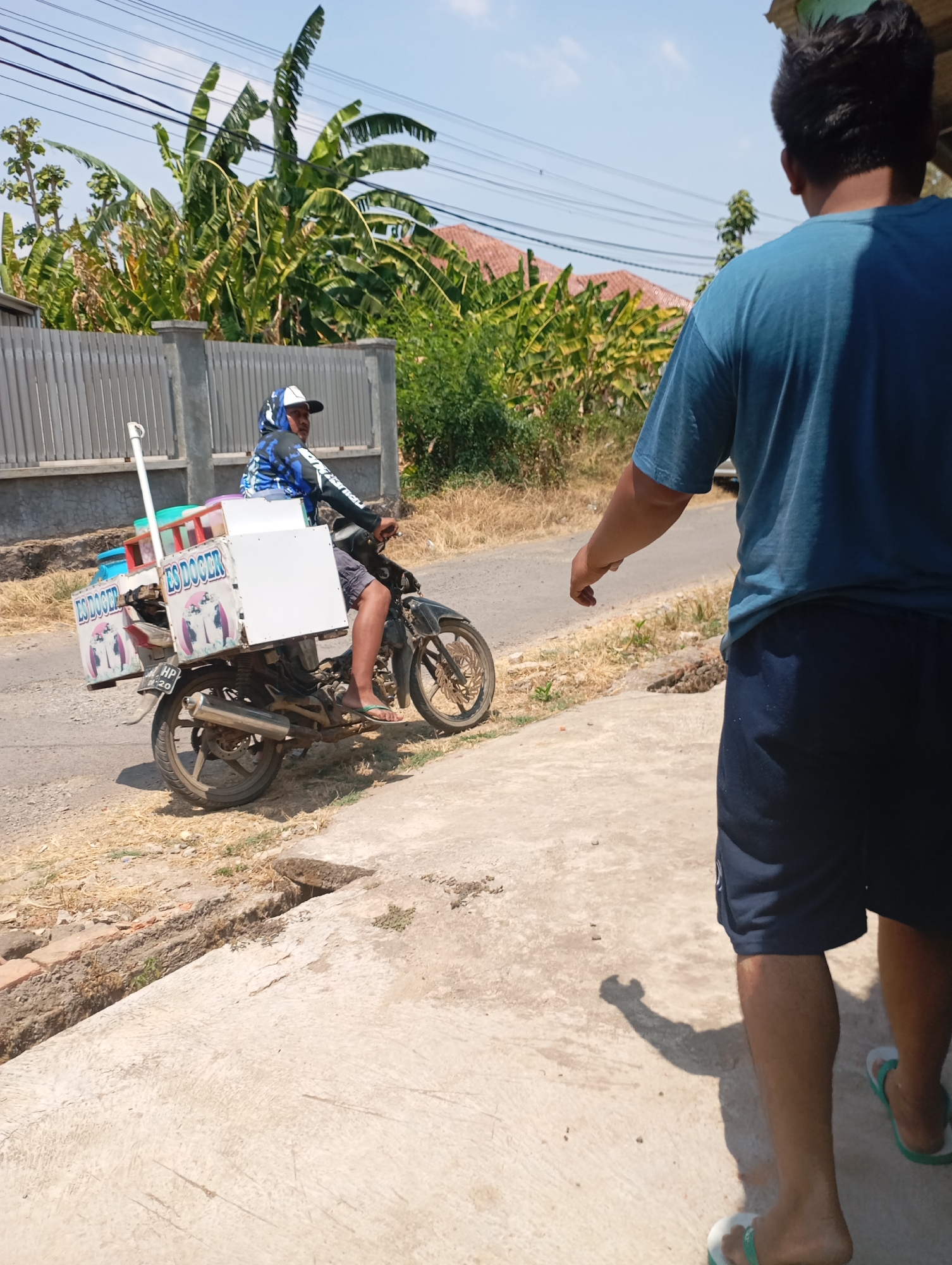 In the foreground, a person is walking away from the camera, seen from the back. They are wearing a blue t-shirt and dark shorts, with white and green flip-flops on their feet. Their shadow is cast on the concrete ground. The environment appears to be a street in Kecamatan Haurgeulis, Indonesia, with dry grass and foliage visible on the side of the road. In the midground, a man is riding a motorcycle that is equipped with a white box on the back, labeled "ES DOGER". He is wearing a blue and black patterned hoodie and a white cap. The motorcycle is parked on the side of the road. Behind the motorcycle, there is a grey fence and lush green banana trees. The sky is clear and blue, indicating a sunny day.