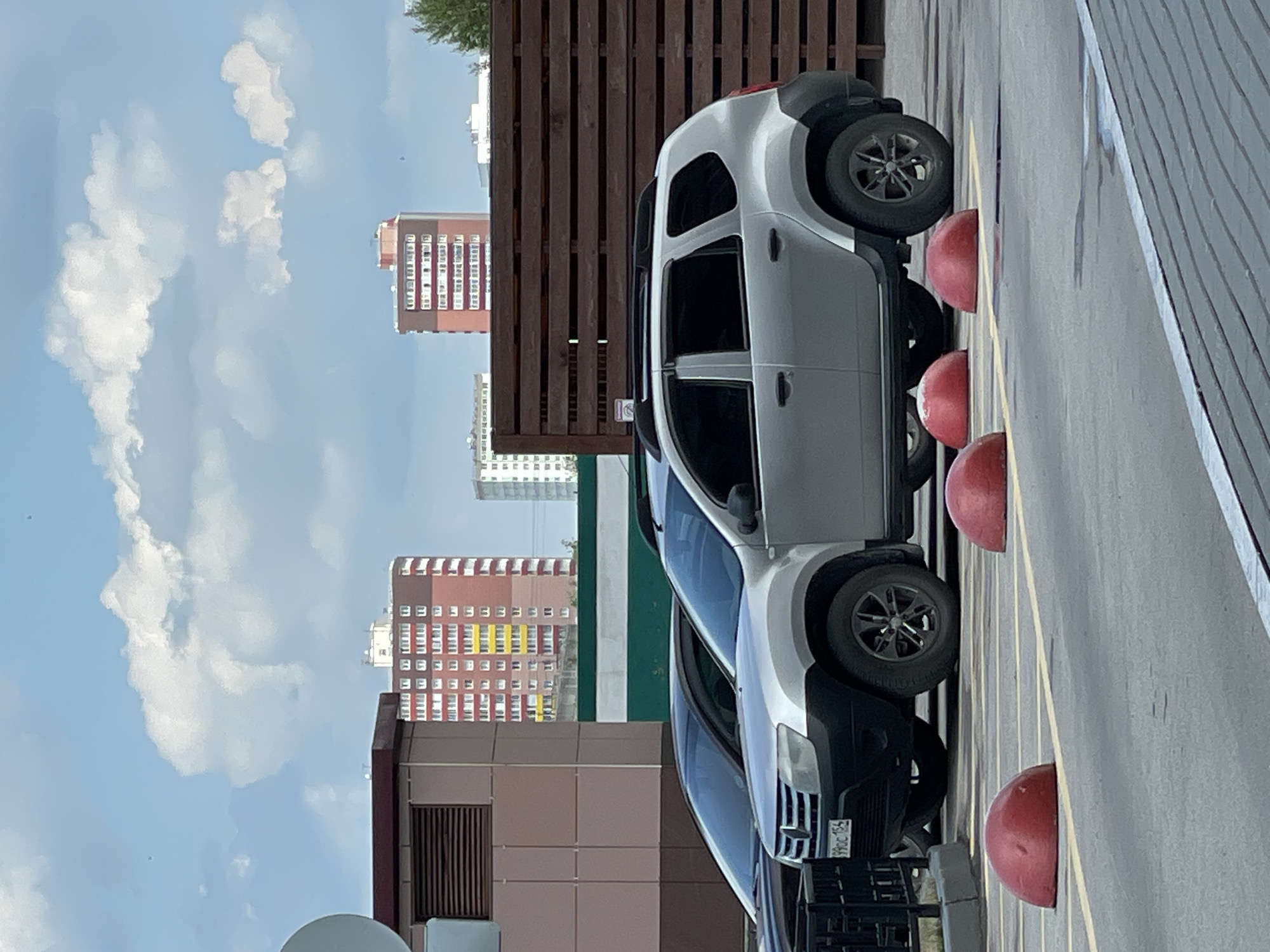 The image captures a scene on a cloudy day in Novosibirsk, Russia, showing a silver Dacia Duster parked on an asphalt surface. The car is in the foreground, facing right, with its front bumper and passenger side visible. A black car is partially visible to the left of the Dacia Duster, parked behind it. In front of the parked cars are three red, dome-shaped bollards, arranged in a line. Yellow road markings are visible on the asphalt.
Behind the cars, a dark brown wooden fence stretches across the mid-ground. Beyond the fence, several buildings are visible. To the left, a large, multi-story building with a brown and beige facade dominates the background. Further to the right, a tall, red brick apartment building with many windows is prominent. Another white building is visible to the right of the red building, and trees are present on the far right edge of the image. The sky is filled with white, cumulus clouds against a blue backdrop. The overall scene appears to be a parking area or street within a city.