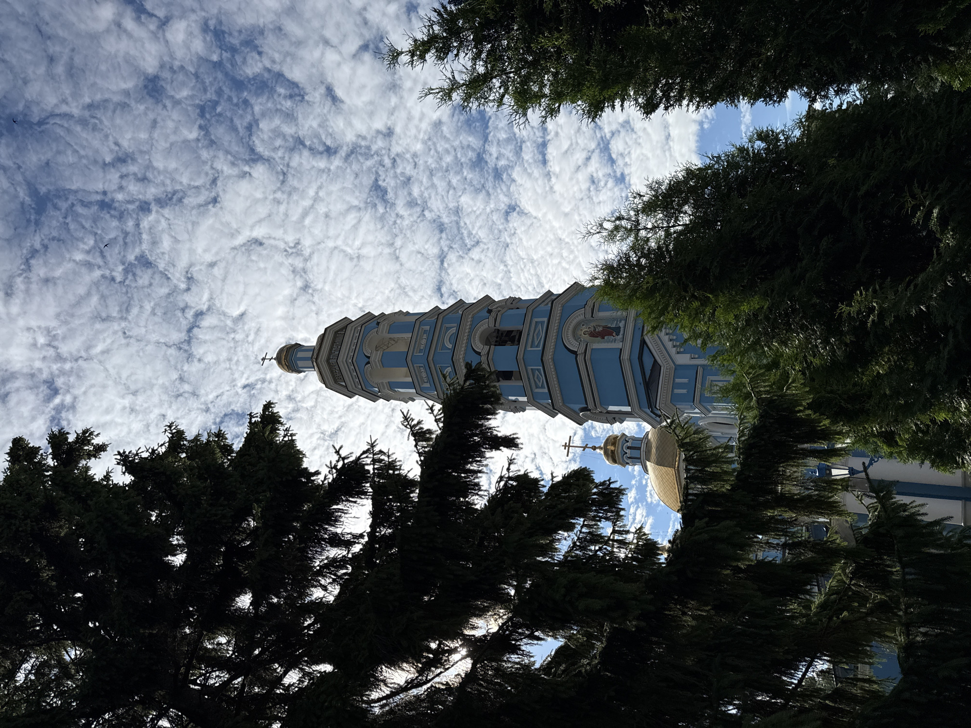 The image shows a bright blue and white bell tower with a gold dome, framed by dark green conifer trees. The tower has multiple levels and is topped with a cross. Above, a dynamic sky filled with white, textured clouds contrasts with patches of blue sky. Two small birds are visible in the distance against the clouds. The overall scene is a view of a religious building peeking through lush greenery under a dramatic sky.