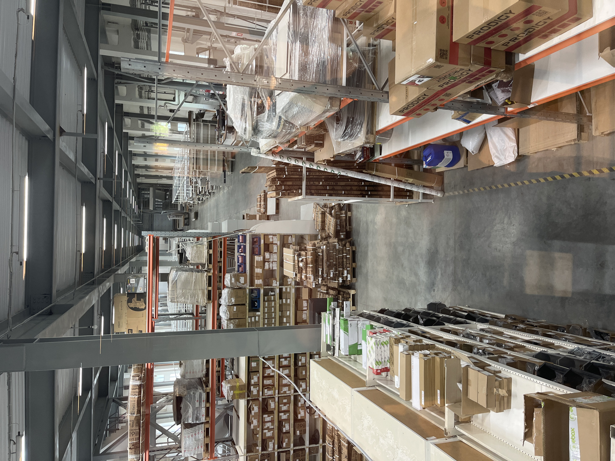 The image presents a wide, elongated interior view of a large, well-lit warehouse or industrial storage facility. Tall metal shelving units extend into the distance along multiple aisles, densely packed with various goods. The shelves are predominantly filled with cardboard boxes of different sizes, many items wrapped in clear plastic, and some products stacked on pallets.
The floor is a plain concrete surface, marked with yellow and black striped safety lines in certain areas. Overhead, the high ceiling is supported by prominent metal structural beams, and the space is brightly illuminated by numerous linear fluorescent light fixtures.
Specific objects visible on the shelves include a blue industrial container and several white bags. Closer details reveal organized smaller items in bins on lower shelves, alongside the larger stacked boxes. No individuals or specific activities are depicted, portraying a static scene of an organized storage environment. The time of day and weather are not discernible due to the indoor setting with artificial lighting.
Visible text includes the brand name "PRODIGY" prominently displayed on numerous cardboard boxes. Other labels read "STALEX" on one larger box and parts of words like "УДОБН" and "ДОМА" (likely forming "УДОБНО ДОМА," meaning "comfortably at home"). The word "ebox" is also discernible on some packaging.