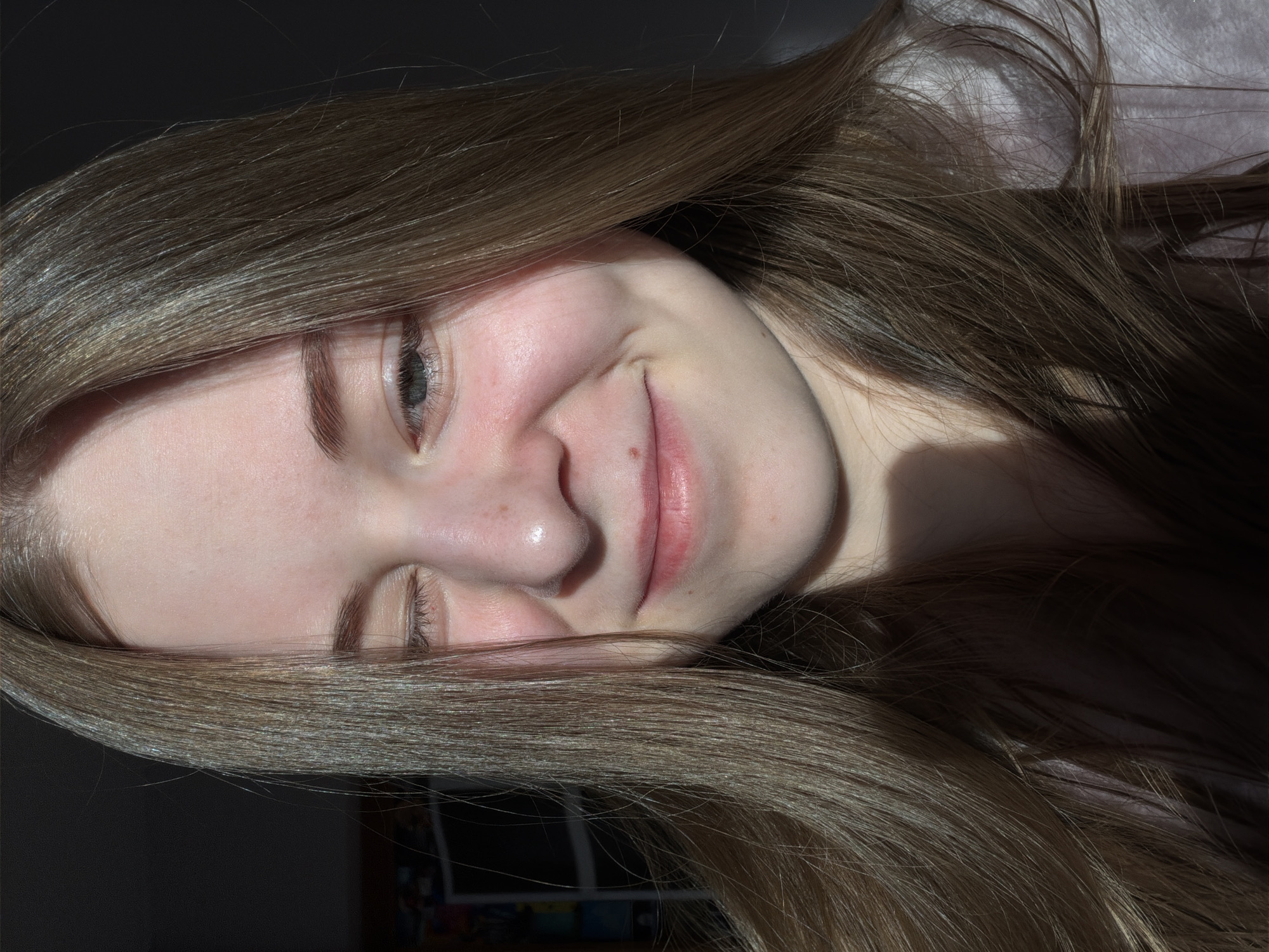 A close-up, slightly angled portrait of a young woman with long, light brown hair. Part of her face is in shadow. Her eyes are partially closed. She is smiling slightly. The background is out of focus and partially dark. There is no discernible information indicating the location in the city of Perm, Russia.
