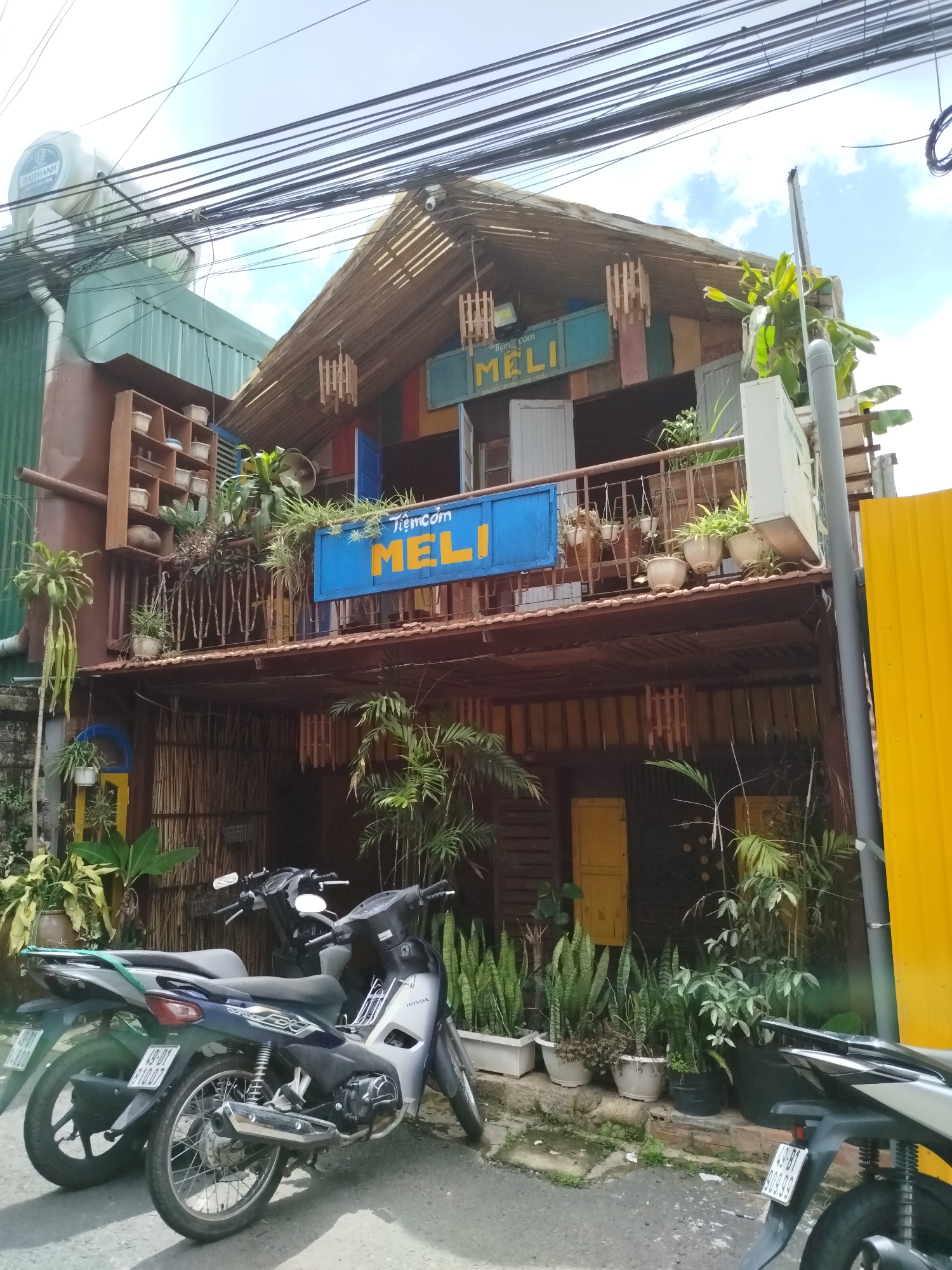 The image shows the exterior of a building in Phường Xuân Hương - Đà Lạt, Vietnam, designed with a rustic, natural aesthetic. The building features a multi-level structure with bamboo and wooden elements. A prominent blue sign with yellow lettering that reads "Tiệm cơm MELI" is displayed on the upper level, indicating it is a cơm (rice) shop. The facade is adorned with numerous potted plants, including snake plants and other tropical foliage, adding to the natural and somewhat overgrown appearance. There are two motorcycles parked in front of the building on a paved area. Above the building, a tangle of electrical wires is visible against a partly cloudy sky. The architecture suggests a casual, possibly bohemian, establishment.