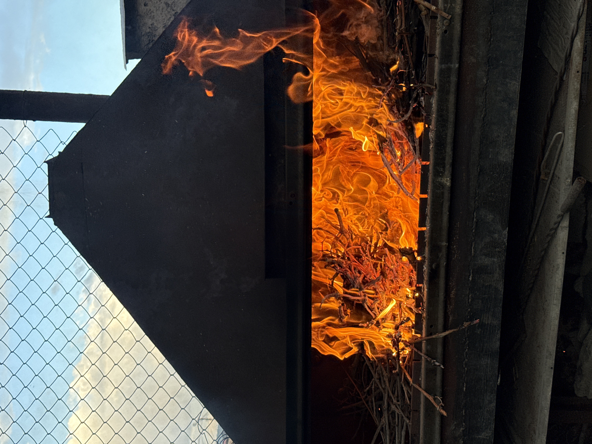 The image shows a fire burning inside or behind what appears to be a metal structure. The flames are orange and yellow, consuming what looks like brush and small branches. To the left of the fire is a dark, angled metal sheet, and beyond that, a wire fence with a sky visible through the mesh. To the right of the fire are metal bars and other structural elements. There is no indication of the location being in Yerevan, Armenia, or the presence of any people.