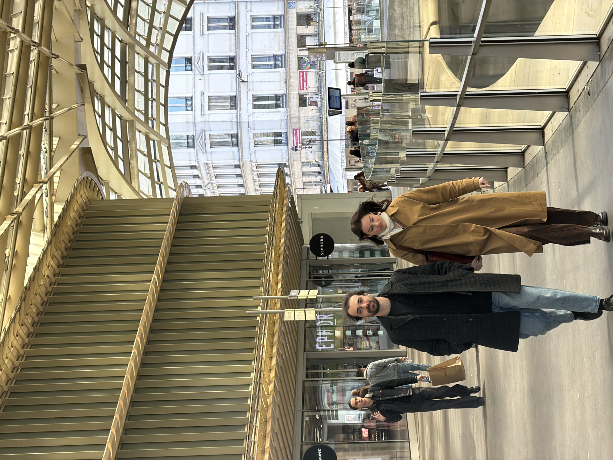 This image depicts a pedestrian area within the Forum des Halles in Paris, France, identifiable by the distinctive golden-hued, undulating Canopée architectural structure overhead and traditional Parisian buildings in the background.
In the foreground, a man and a woman walk side-by-side towards the viewer, holding hands. The man is Caucasian, with dark curly hair and a beard, wearing a dark grey textured overcoat, a dark t-shirt, light blue jeans, and dark shoes. He is looking directly at the camera with a slight smile. The woman is Caucasian, with dark hair slightly wind-blown, wearing a light brown trench coat, a white high-neck top or scarf, dark brown ribbed trousers, and dark brown shoes. She carries a dark red shoulder bag on her left shoulder and looks slightly downwards and to her right.
Behind them on the left, a man in a dark jacket and trousers walks away from the camera, carrying a brown paper shopping bag labeled "LOUE". Further back, another person in a denim jacket and jeans is visible. The ground is a light grey paved surface. To the right, a modern glass and metal railing, featuring distinct vertical silver posts, borders the pedestrian path.
On the left side of the scene, retail storefronts are visible through large glass panels, including a prominent "SEPHORA" sign and another store partially labeled "SKINE". Overhead, the modern, golden Canopée structure spans the scene, characterized by its intricate metal framework and translucent panels. In the background, above and behind the Canopée, a multi-story, white classical Parisian building with numerous windows, some featuring decorative balconies, is visible. Below this building, additional pedestrians, street furniture, and commercial facades are discernible, indicating an active urban setting under natural daylight.