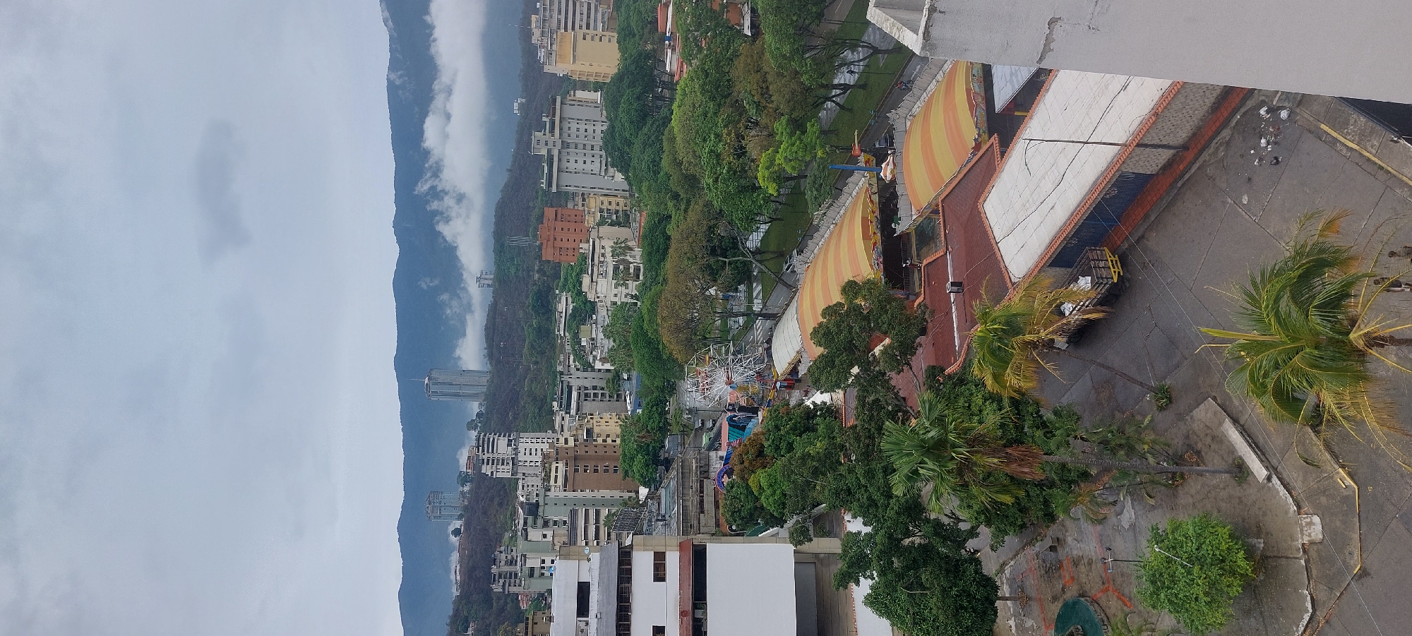 This is an elevated view of Caracas, Venezuela, showing a cityscape nestled against a mountain range obscured by heavy clouds. The built environment consists of closely packed, mostly mid-rise buildings. Vegetation is dense and widespread. Near the foreground is an amusement park with a Ferris wheel and striped tent-like structures. In the immediate foreground, there are palm trees and concrete areas with street furniture, possibly a plaza. The weather is overcast. A forensic description is limited by the distance and angle of the shot, making it difficult to discern details about people, interactions, or the precise function of objects.