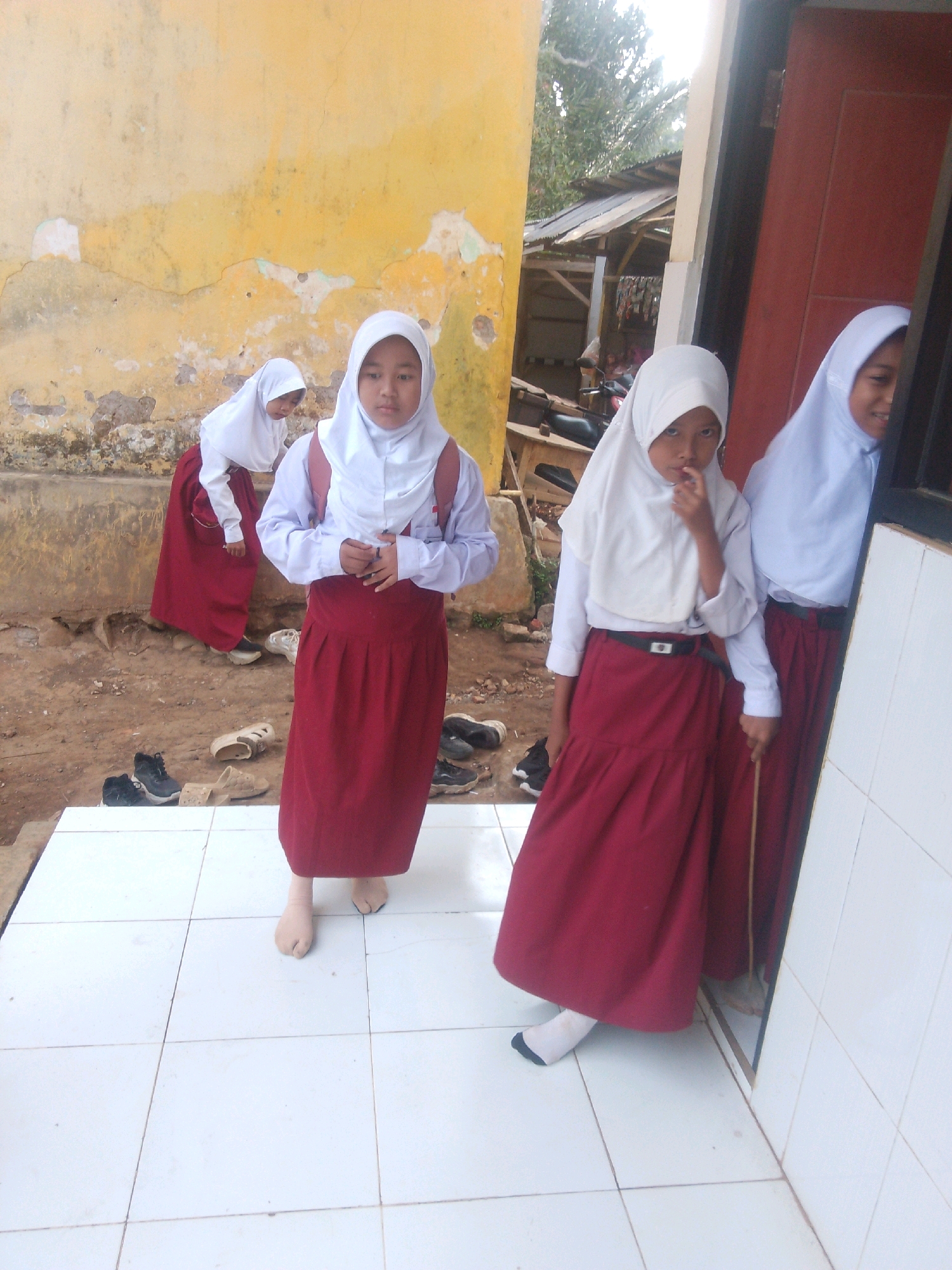 Four young girls in school uniforms are standing outside a building. The girl in the foreground, facing the camera, wears a white hijab and a long maroon skirt. She has her right hand to her mouth. Behind her and to her left, another girl also wears a white hijab and maroon skirt, looking down. To the right of the foreground girl, partially obscured by a doorway, stands a third girl in a white hijab and maroon skirt. Further left and in the background, a fourth girl in a white hijab and maroon skirt is visible. The building has a weathered yellow wall and a reddish-brown door. The ground in front of the building is a tiled patio, with dirt and scattered shoes visible in the background. Some trees and a small structure with a corrugated roof are also present in the background. The scene appears to be an outdoor school environment in Betek, Indonesia.