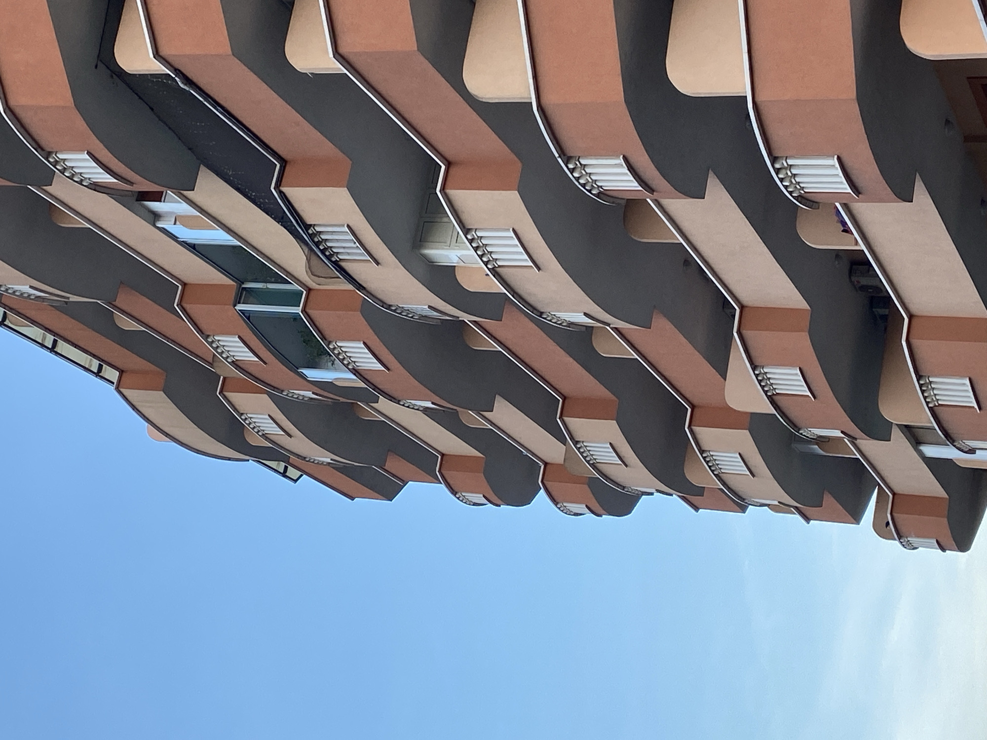 The image shows a low-angle view of a building facade in Budva, Montenegro. The architecture features horizontally layered, wave-like extensions painted in alternating bands of brown, dark gray, and beige. Each extension has small windows or vents with visible white horizontal slats. The background is a clear blue sky. The photograph seems to have been taken looking upward, emphasizing the building's overhanging design.