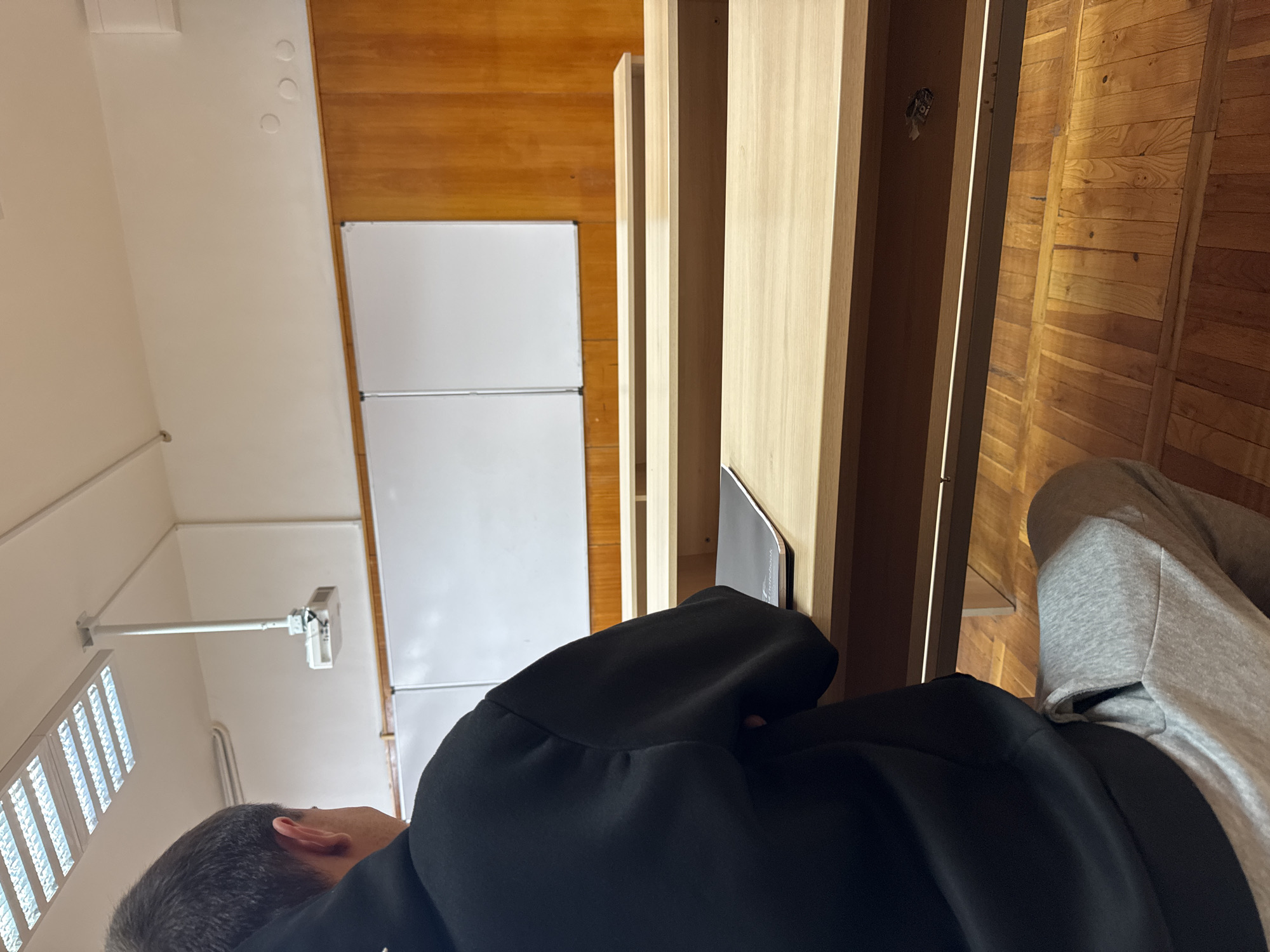 An interior image depicts a classroom or lecture hall in Varna, Bulgaria, viewed from the perspective of a seated individual. The primary subject is the back of a male individual, presumably a student, seated in the foreground. He possesses dark hair, wears a black hooded sweatshirt, and grey sweatpants. His hands are out of view, and a black notebook or binder rests on the light-colored wooden desk directly in front of him.
The room features multiple rows of light-colored wooden desks and benches, arranged in an ascending manner consistent with a lecture hall layout. The floor is composed of light-colored wooden planks. The front of the room displays two rectangular whiteboards mounted against a wall. Above the whiteboards, the wall transitions to wood paneling, while the upper section of the room walls is painted white.
Overhead, multiple rectangular fluorescent light fixtures illuminate the space. A white digital projector is suspended from the ceiling by a pole, positioned in front of the light fixtures and above the whiteboards. The overall environment is academic, orderly, and well-lit.