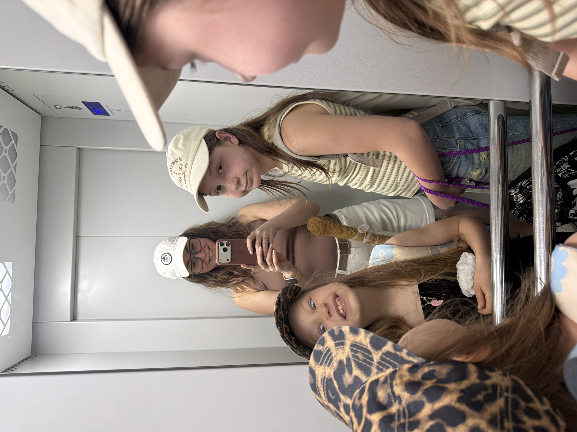 Four young individuals, likely girls, are taking a selfie in an elevator. The elevator's interior is a simple white. A girl in a white hat and glasses is taking the photo with a smartphone. Other girls are partially visible in the reflection, one wearing a leopard-print hat, and another in a striped shirt and hat. The specific location in Chaikovsky, Russia, is not discernible from the image.