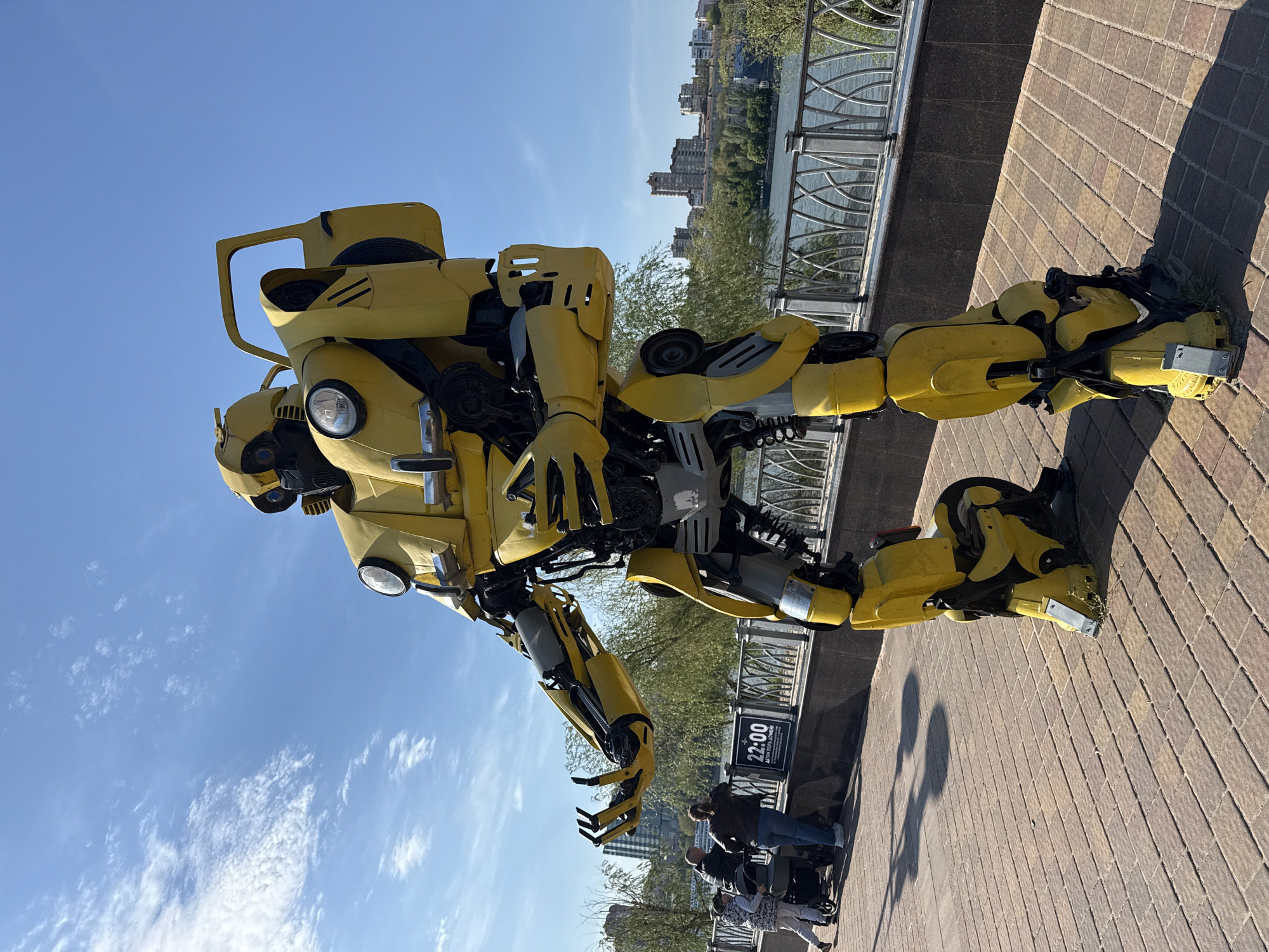 The image shows a yellow robot sculpture, resembling the Transformer Bumblebee, positioned outdoors in Rostov-on-Don, Russia. It stands on a brick pathway adjacent to a metal railing overlooking a body of water and buildings in the background. The sky is blue with scattered clouds. In the lower right, people are standing or sitting next to the railing. A sign with the time "22:00" is visible near the group.