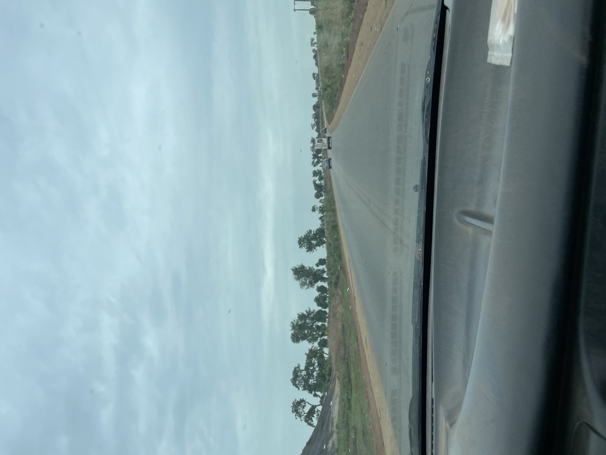 View from inside a vehicle traveling on a paved road lined with trees in Kujama, Nigeria. The sky is overcast. A few vehicles are visible in the distance ahead. The vehicle's interior and windshield are partially visible.