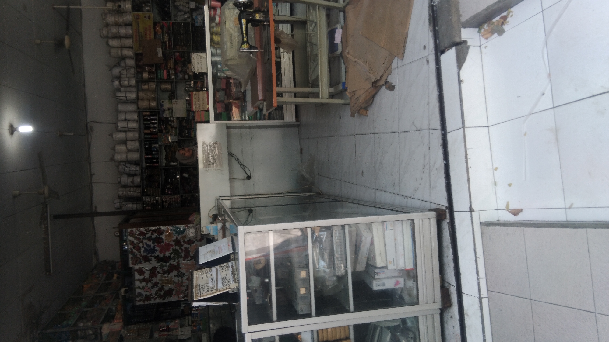 A medium shot captures the interior of a shop, likely a fabric or sewing supply store, in Pekalongan, Indonesia. The shop appears to be on street level with the entrance open to the outside, as suggested by the tiling that transitions from inside to outside.
The shop is filled with shelves stacked high with various goods. On the left, a glass display cabinet showcases items on multiple shelves, including what appear to be electronic devices or equipment, and boxed goods. To the right of the display cabinet, a counter area is visible, with a woman wearing a hijab looking towards the right of the frame. Behind the counter, shelves are packed with items that resemble spools of thread or similar materials. Further to the right, a sewing machine sits on a wooden table, covered by a tarp.
The overall environment is dimly lit, with overhead lighting visible. The flooring is tiled throughout the interior, with some of the tiles outside the shop appearing weathered or broken. There are also stacks of brown cardboard boxes or fabric on the floor near the sewing machine. The shop displays some signs, including a price list on the left, which is partially obscured. The overall impression is of a functional, cluttered retail space.