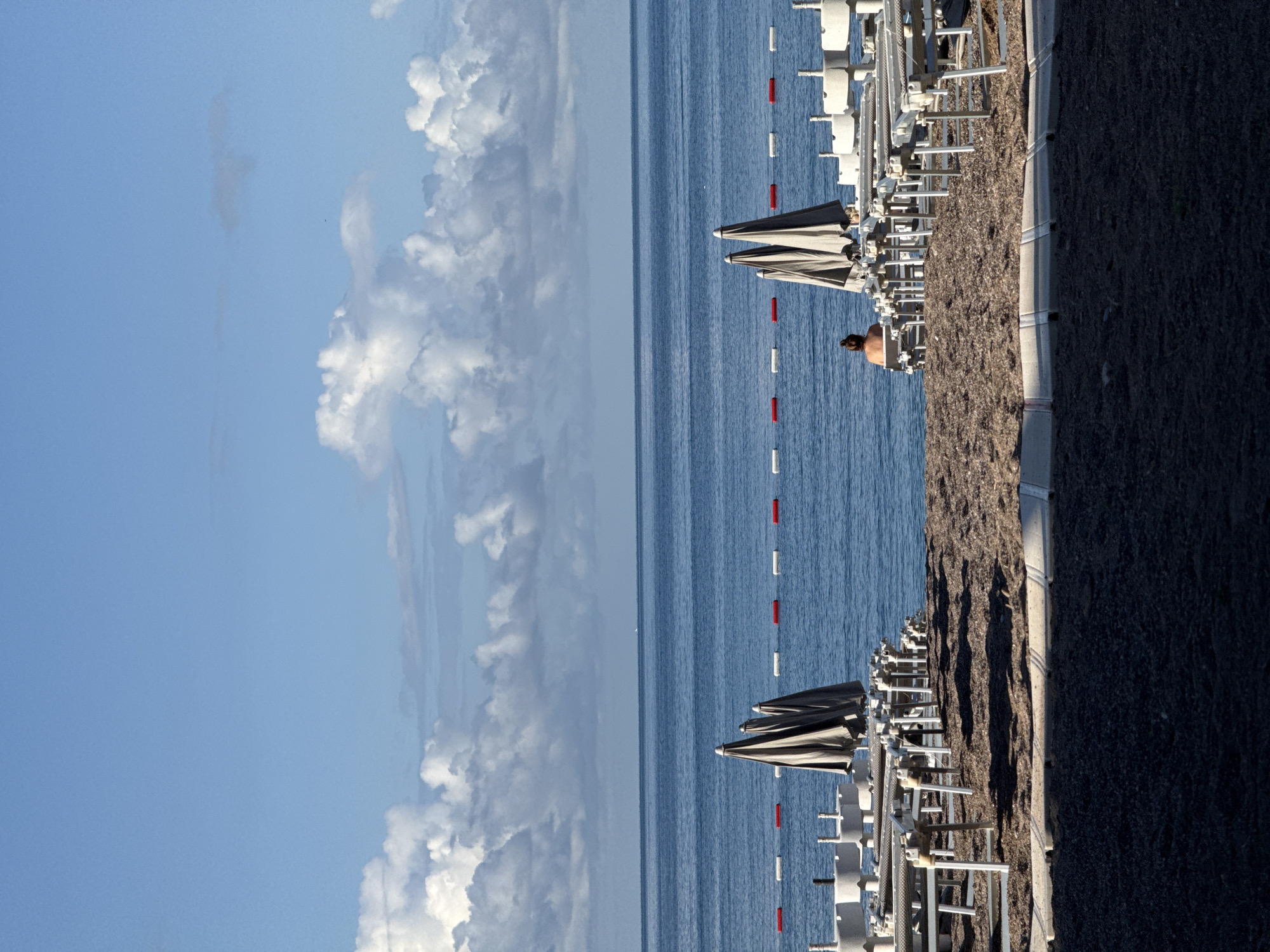 The image shows a beach in Rafailovići, Montenegro, with a view of the Adriatic Sea and the sky. The beach is lined with rows of sunbeds and closed umbrellas. A person with their back towards the camera is standing on the beach near the sunbeds. The sea is calm with a few red and white buoys floating on the surface. The sky is blue with scattered white clouds. A concrete pathway separates the beach from the shoreline.