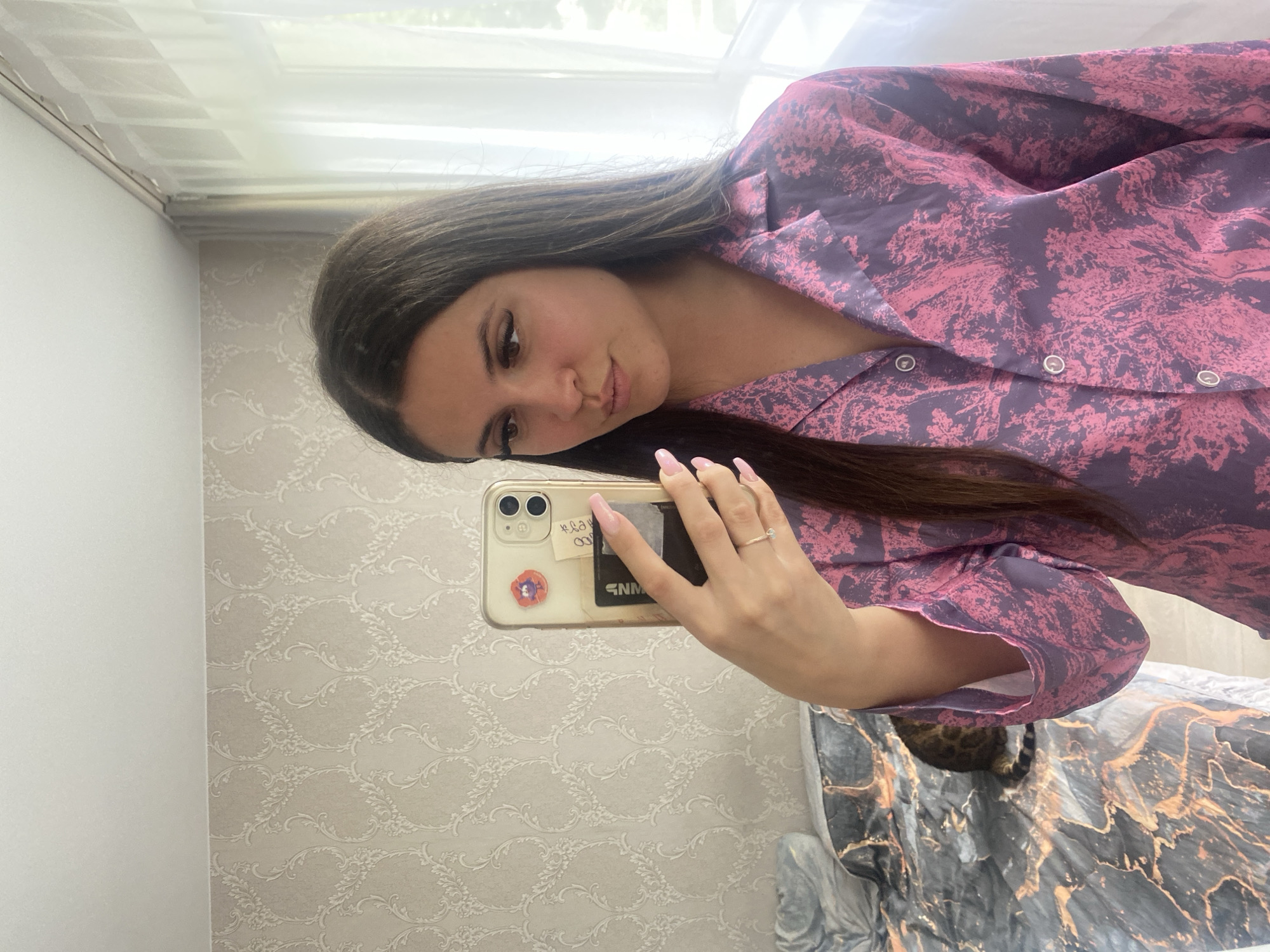A young woman is depicted taking a mirror selfie in what is described as a residence located in Chaikovsky, Russia. She has long, straight dark brown hair and is wearing a long-sleeved, collared button-up shirt featuring a dark purple background with an intricate, dense pattern of bright pink botanical or abstract tree-like motifs. Her nails are painted light pink, and she wears a delicate ring with a small, clear stone on her right ring finger. She holds a light-colored smartphone with dual prominent cameras in a clear case, which partially obscures her face and upper body as she looks towards the screen with slightly pursed lips. The phone case has a small red poppy-like sticker and what appear to be cards or notes tucked into the back.
The background features a light beige wall adorned with a repeating, textured damask wallpaper pattern in off-white or light gray. To the right of the subject, a large window allows bright natural light into the room, diffused by sheer white curtains that hang from a visible curtain rod. Green foliage is faintly visible through the window. On the lower left of the frame, a bed is partially visible, covered with a duvet featuring a dark gray and black marble-like pattern with prominent swirling coppery-gold or orange accents. A cat, with dark fur and visible stripes on its tail, is curled on the bed, mostly obscured by the woman's arm. The ceiling above is plain white.