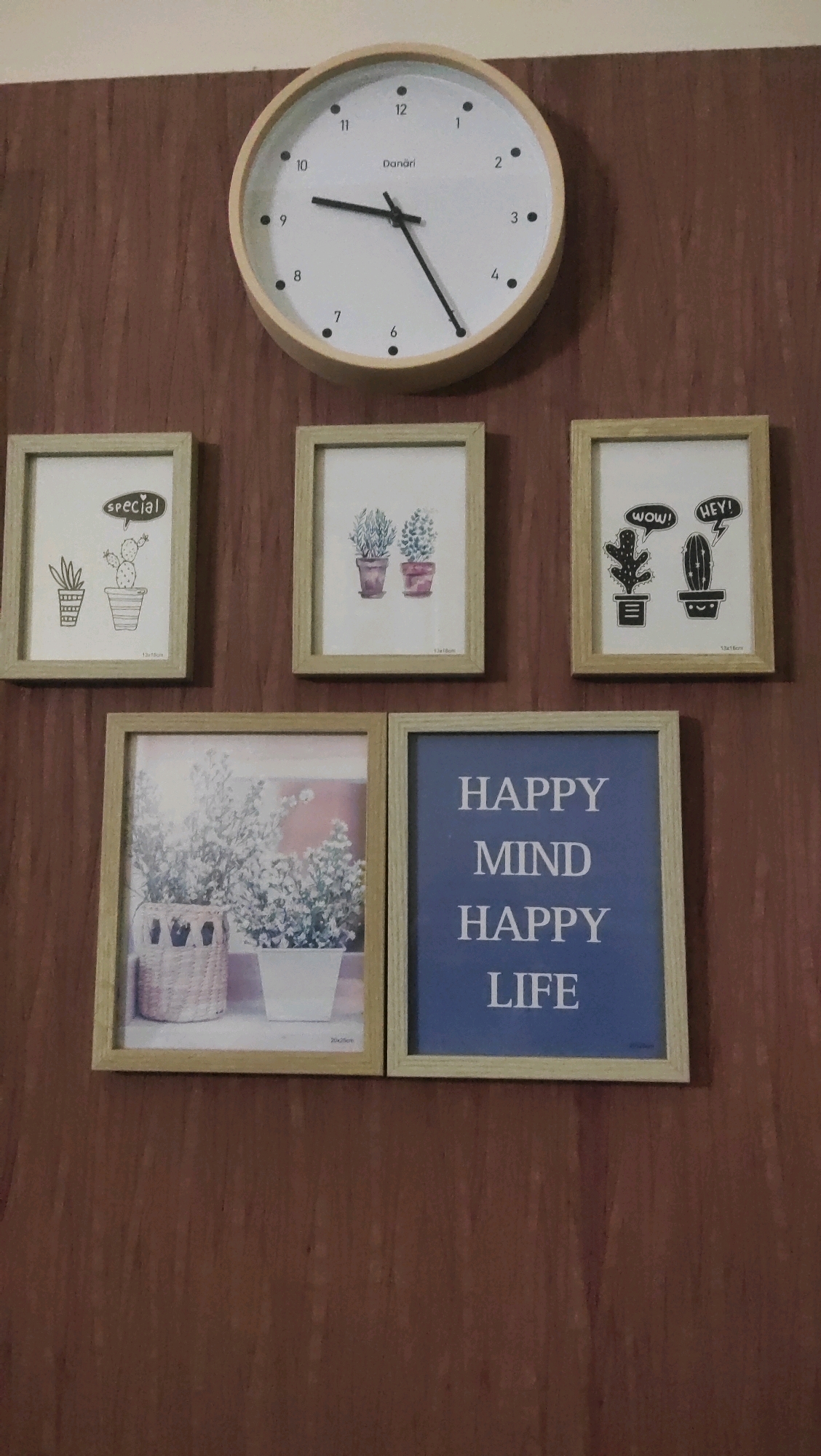 A round wall clock with a light wood frame is centrally positioned above a collection of framed pictures, all mounted on a wood-paneled wall. The clock face features black hour markers and a Danori logo. The pictures are arranged in two rows: three small square frames in the top row and two larger rectangular frames in the bottom row. The top-left frame contains a drawing of potted plants and the word "special" in a speech bubble. The middle frame shows two potted plants. The top-right frame features cartoonish drawings of cacti with speech bubbles containing "WOW!" and "HEY!". The bottom-left frame displays a photograph of potted plants in woven and white planters. The bottom-right frame contains a blue background with white text that reads "HAPPY MIND HAPPY LIFE". The scene is indoors, suggesting a home or office environment.