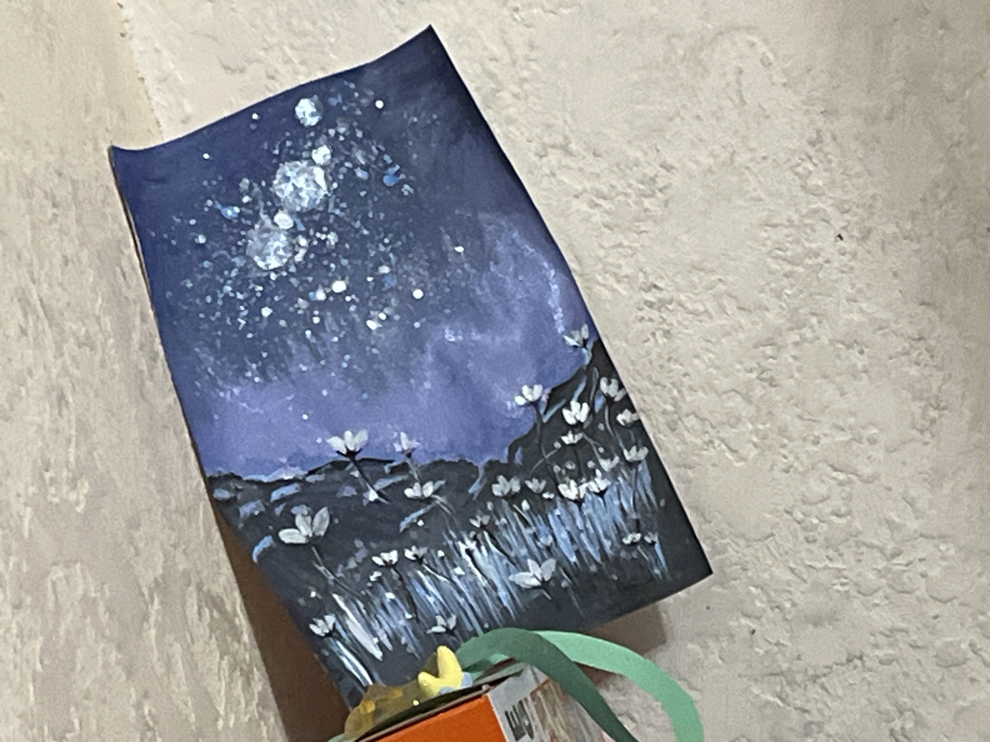 A close-up, high-angle shot shows a piece of artwork leaning against a textured surface, possibly a wall or floor. The artwork is a dark blue painting depicting a starry night sky with a cluster of bright, white celestial bodies at the top. Below the sky is a dark landscape with stylized white flowers and green grass. The painting appears to be on a rigid surface, possibly paper or canvas, which is bent slightly at the top left corner. A small portion of an orange box and green ribbon are visible at the bottom, suggesting the artwork may be part of a gift or decoration. The background is a light-colored, textured surface with an irregular, mottled pattern. There are no people or animals visible in the frame. The location of Krasnyye Barrikady, Russia is not indicated by any visual elements in the image.