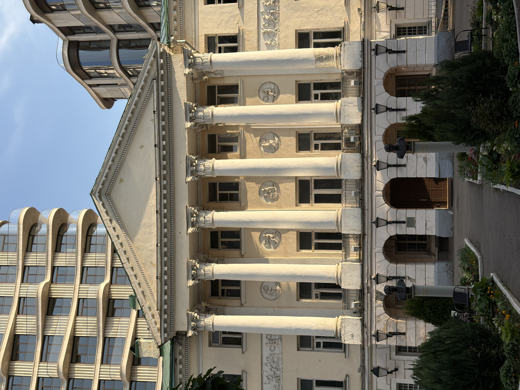 The image captures a view of the Moscow Architectural Institute (MARCHI) building in Moscow, Russia. The building exhibits classical architectural features, including white columns, a pediment, and decorative reliefs. Windows are visible between the columns. Adjacent to the MARCHI building is a more modern structure with a curved facade and multiple windows. In front of the MARCHI building, there is a walkway, a bust on a pedestal, lamp posts, and greenery. The sky is visible above the buildings, suggesting daylight.