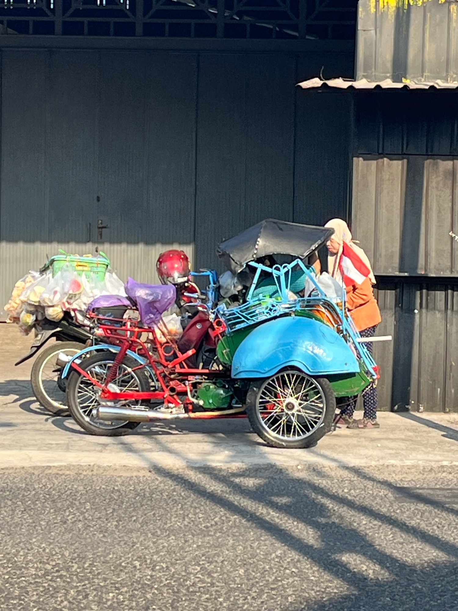 In Yogyakarta, Indonesia, a person wearing a headscarf, orange jacket, and patterned pants stands next to a bright blue and green pedicab parked on a paved surface in front of a gray metal wall. The pedicab, which appears to be custom-built on a motorcycle frame, is laden with various items, including a green bin filled with plastic bags, a red helmet, and other bundled goods covered in plastic. A small Indonesian flag is attached to the pedicab's side. The sun casts long shadows across the ground, indicating it is likely daytime. The background consists of a large, corrugated metal structure with a darker section above it.