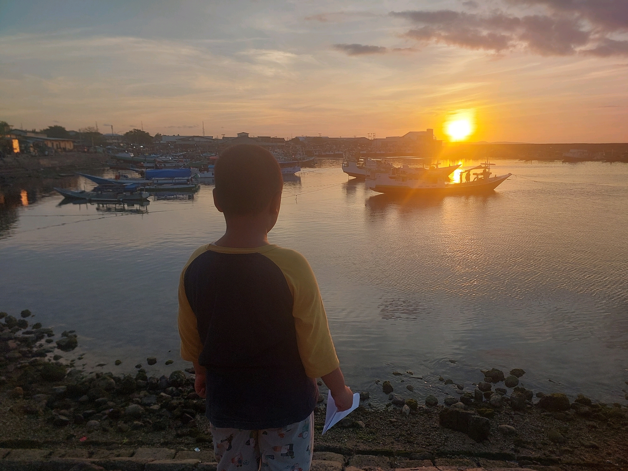 A young child stands on a rocky shoreline facing a body of water with several boats docked or anchored. The sun is setting on the horizon, casting a warm orange glow across the sky and water. In the background, buildings and infrastructure are visible along the coastline. The scene appears to be a harbor or port area in Kupang, Indonesia, given the visual context of the boats and the architecture. The child is holding a paper airplane in their right hand. The water is calm with gentle ripples, and the sky is filled with soft clouds reflecting the sunset.