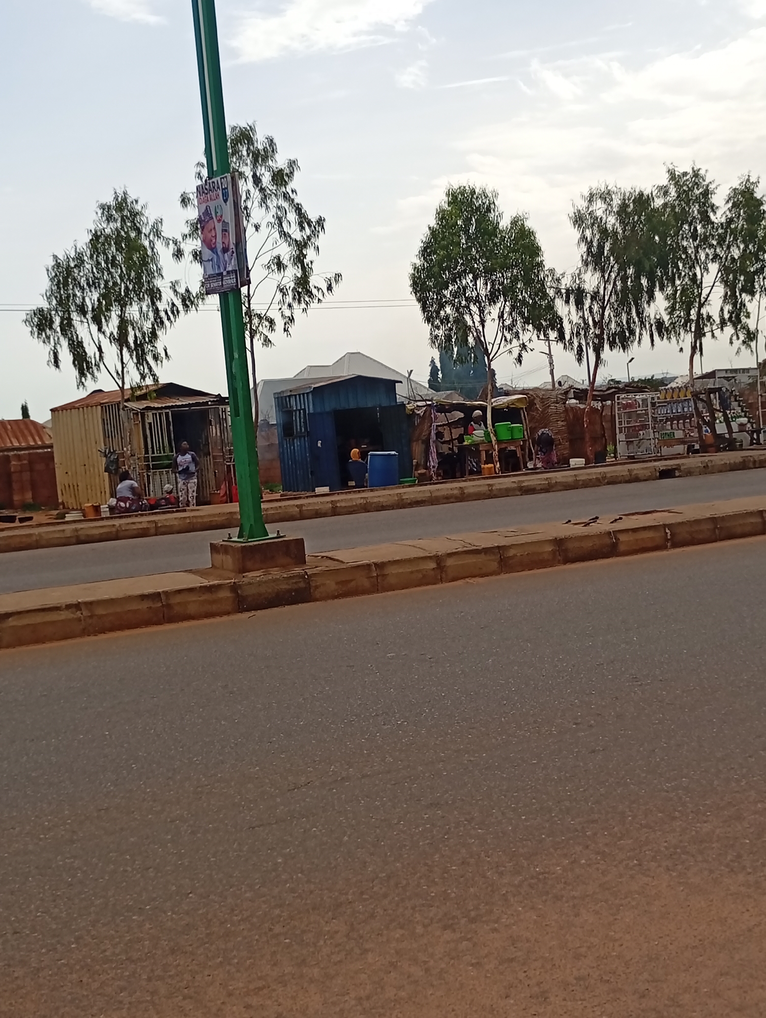 A street-level, medium shot captures a scene in the city of Wuro Dole, Nigeria, featuring a roadside market with several stalls and a streetlamp. The foreground is dominated by a paved road and a concrete curb. In the background, several temporary structures, likely market stalls, are visible along the side of the road. One stall appears to be made from corrugated metal, while another is a blue shipping container converted into a shop. Various goods, including bottles and containers, are displayed on shelves outside some of the stalls. Trees with thin trunks and sparse foliage line the area behind the stalls. A tall, green streetlamp stands prominently to the left of the frame, displaying a political poster with images of two men and text. Several individuals are present, some seen standing or sitting near the stalls, suggesting commercial activity. The sky above is overcast with scattered clouds.