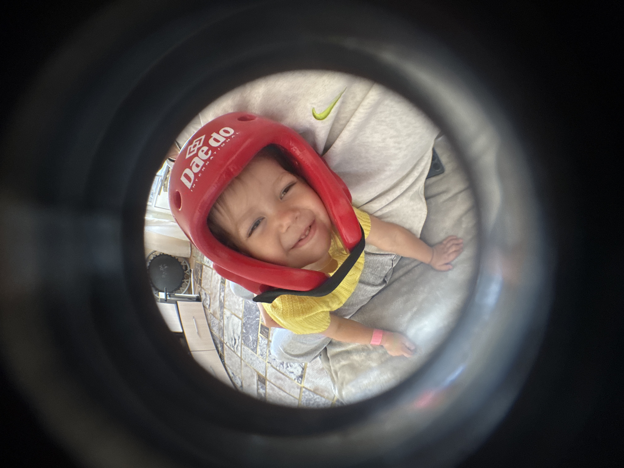 A fisheye-style photograph captures a young child, likely a toddler, wearing a red protective sports helmet. The child is looking upwards with a wide, joyful smile, appearing happy and content. The red helmet has "Dae do" and "INTERNATIONAL" written in white text on its side. The child is dressed in a yellow short-sleeved top and light grey pants, and a pink wristband is visible on their right wrist.
The child is being held or is sitting on an adult's lap, as evidenced by a leg clad in light grey pants with a green Nike swoosh logo visible in the background. The indoor setting features a tiled floor with a patterned design. To the left, a portion of a small trampoline or similar exercise equipment with a black mat and metal frame can be seen. The image is brightly lit, indicating daytime. The strong circular vignette around the central subject gives the impression of a view through a peephole or a unique lens. The scene depicts a playful moment, possibly during or after a supervised activity, taking place indoors in Chaikovsky, Russia.