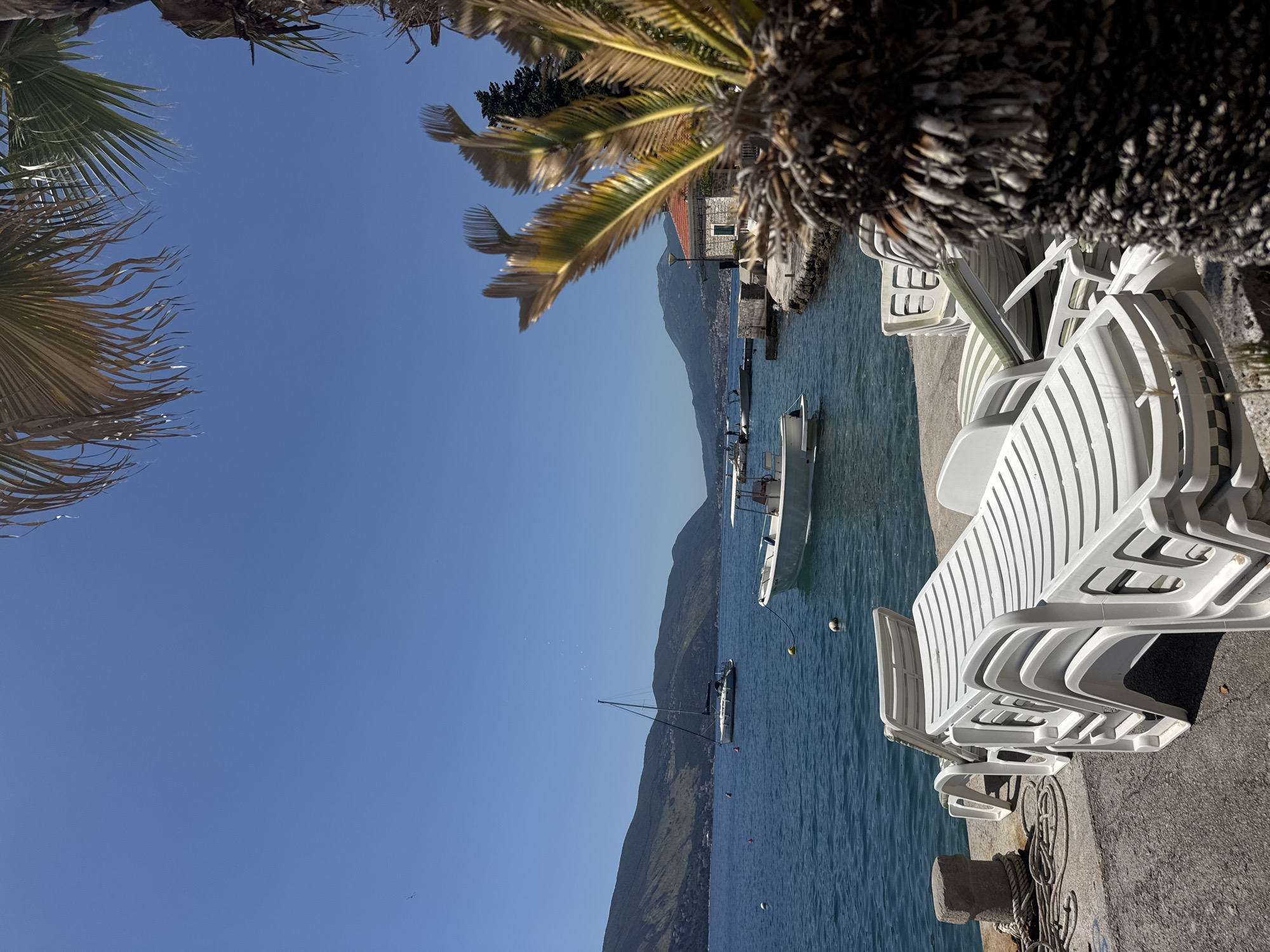 The image displays a sunlit coastal scene in the city of Unknown, Unknown. A clear, cloudless blue sky occupies the upper portion of the frame. In the midground, a calm, deep blue body of water features multiple boats. A white sailboat with a single mast is anchored in the left-center, while a white motorboat with an open canopy is positioned towards the right foreground. Several orange and white buoys are visible on the water's surface. The background consists of a range of green, vegetated mountains extending across the horizon. On the right shoreline, a small, light-colored stone building with a red tiled roof stands on a concrete embankment. The immediate foreground on the left includes a concrete pier with a grey bollard, around which thick ropes are coiled. Stacked on this pier are five white plastic sun loungers. One additional sun lounger with a green and white striped cushion is partially visible behind the stack, in a reclined position. The far right foreground is dominated by the blurred, textured trunk and fronds of a palm tree, which partially frames the scene. No individuals are visible.