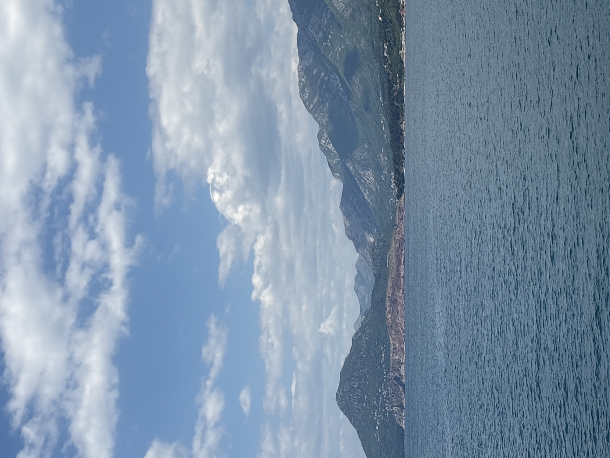 The image shows a view of the Adriatic Sea with a mountainous coastline, likely in the vicinity of Bar, Montenegro. The sky is partially covered with white clouds against a blue backdrop. The water surface appears to be slightly choppy. A series of mountains or cliffs with varying shades of green and brown are visible along the coastline.
