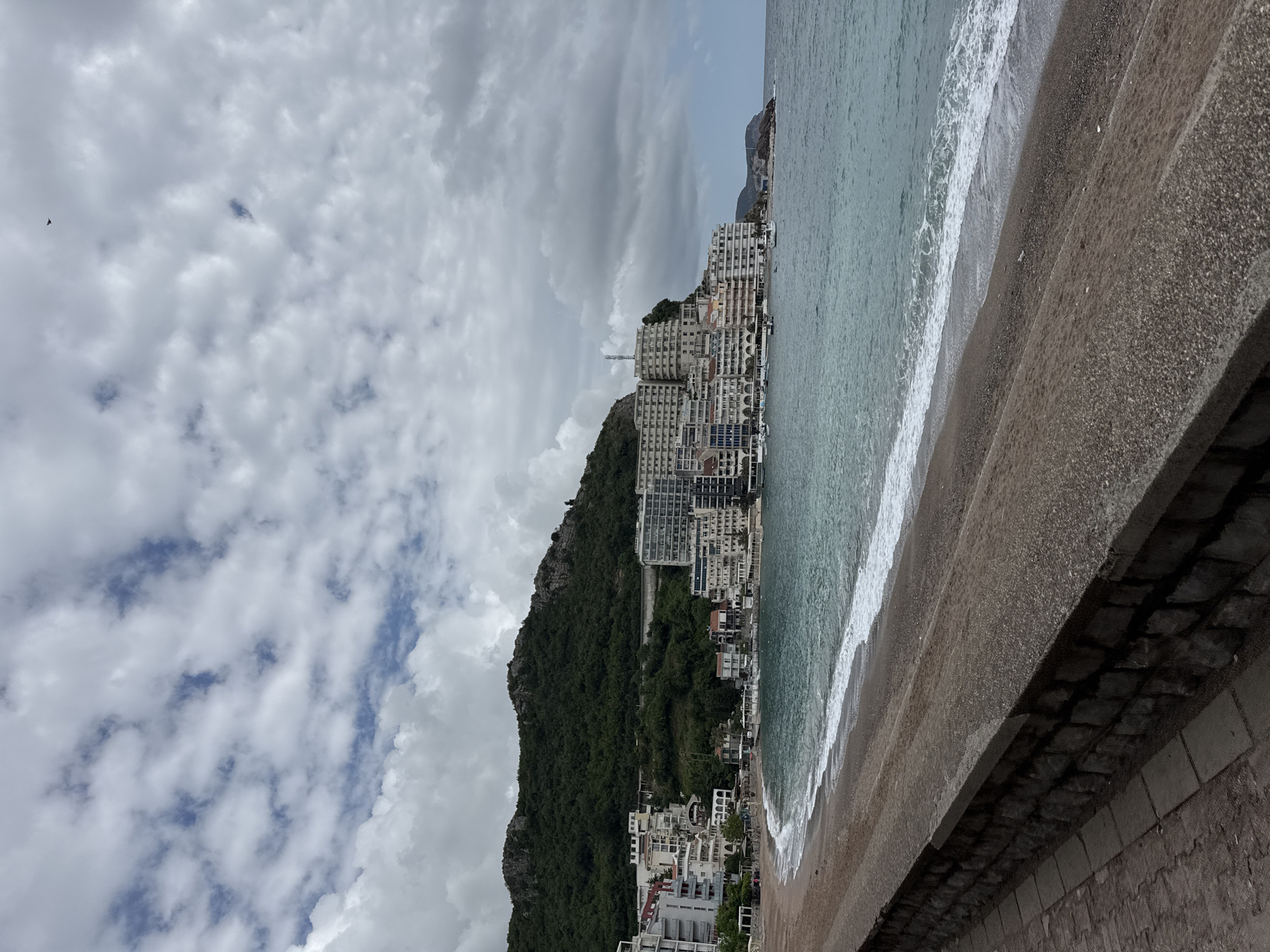The image shows a coastal scene in Rafailovići, Montenegro, with a beach and a cliffside densely built with multi-story buildings. The sea is on the right, with waves breaking on the dark sand. A concrete wall runs along the bottom of the frame, separating the viewer from the beach. Above, a steep, wooded hill supports a vertical arrangement of apartments and hotels, characteristic of the town's development against the natural terrain. The sky is overcast with thick, patchy clouds filling the upper portion of the frame. No people are discernible in the landscape.