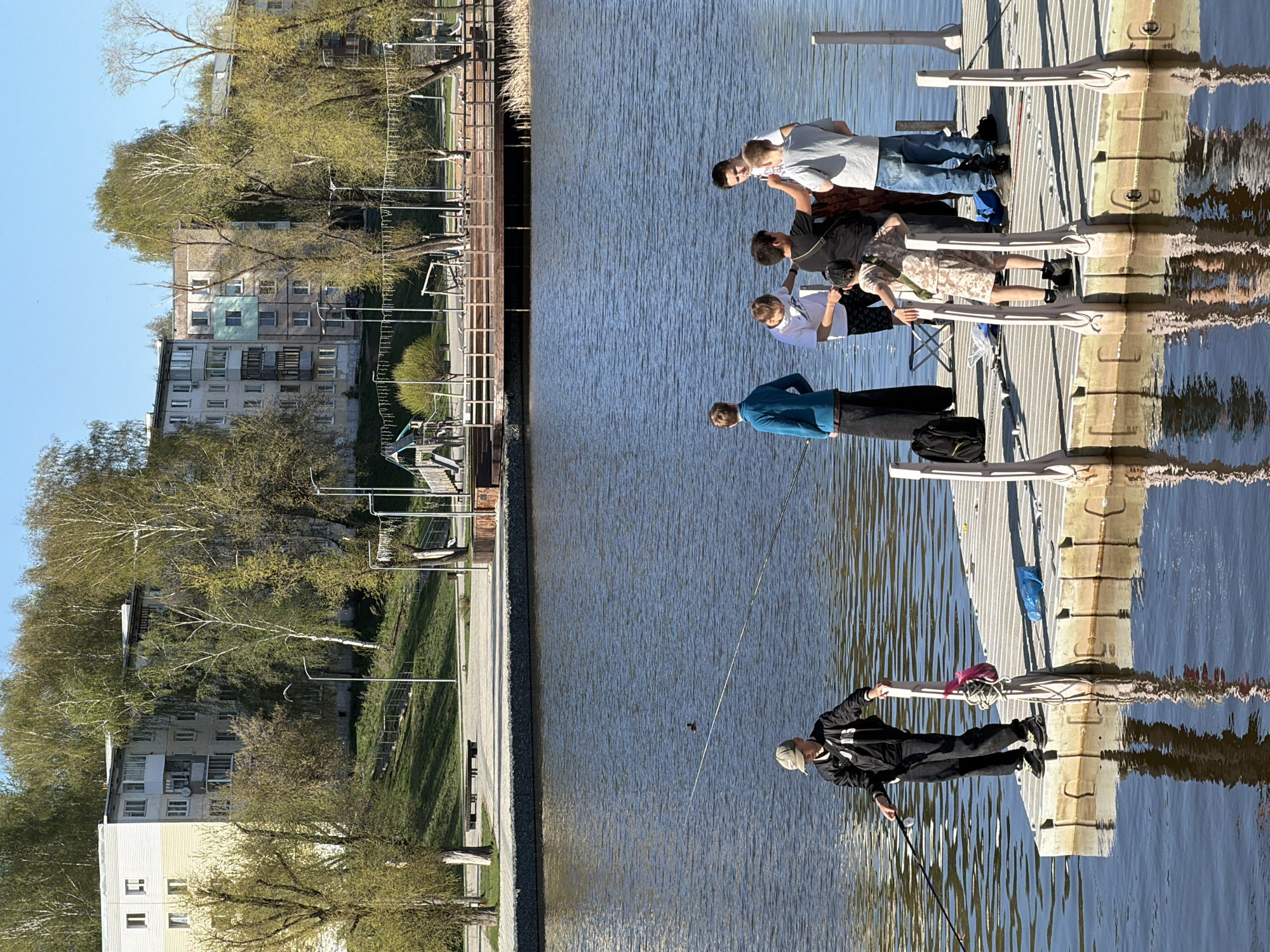 The image depicts a group of people on a floating dock on a body of water, potentially a lake or river, in Votkinsk, Russia. Some are fishing, while others appear to be observing or socializing. The background shows a building and a park area with trees and a walkway. The weather appears sunny.