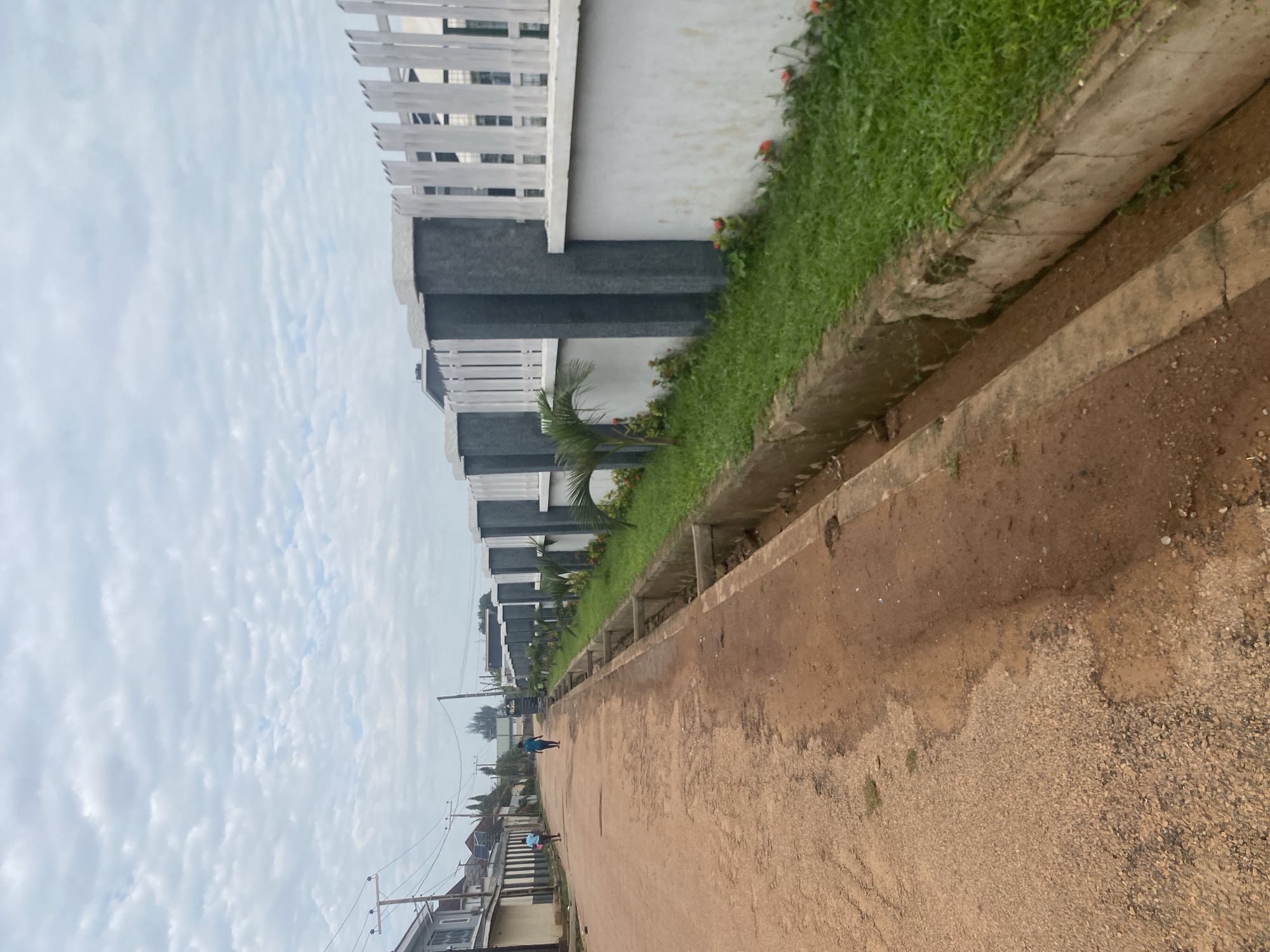 The image shows a street scene in Barden Yana, Nigeria. A dirt road runs diagonally from the foreground to the background, with a concrete drainage ditch running alongside it. On the right side of the road, there is a white wall with grey pillars topped with white vertical planks, suggesting a fence or boundary. A strip of green grass with small red flowers is planted between the drainage ditch and the wall. Several small palm saplings are also visible in this grassy area. In the background, on the left side of the road, several buildings with varying architectural styles are visible. Two individuals are walking away from the camera on the road. The sky above is overcast with scattered clouds.