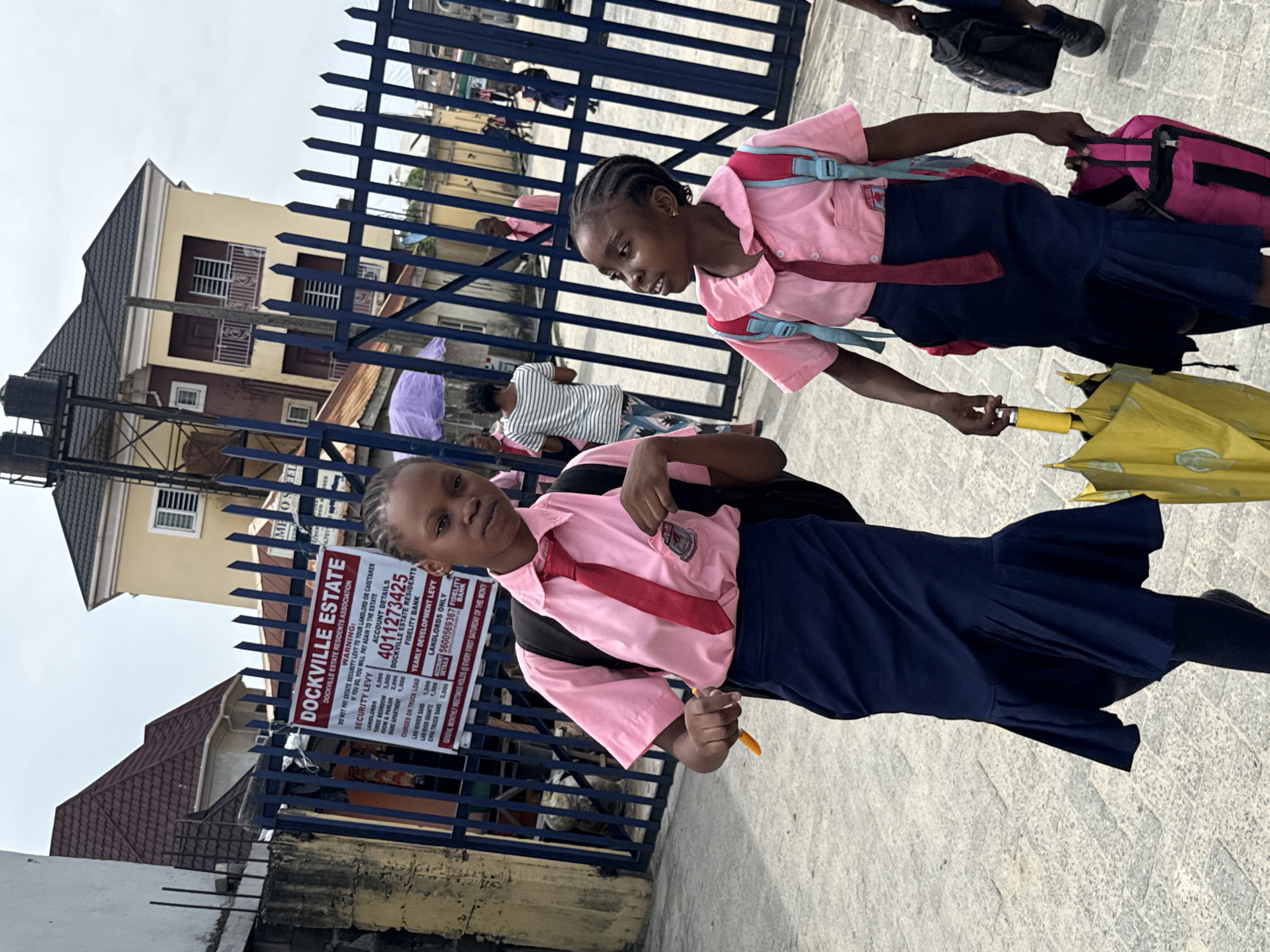 Two young girls in pink school uniforms and navy blue skirts walk on a paved surface, likely a sidewalk, in front of a blue metal fence. The girl in the foreground holds a yellow object, possibly an umbrella. A sign on the fence reads "DOCKVILLE ESTATE", providing instructions for security levy payments and listing a Fidelity Bank account number. Buildings are visible behind the fence, suggesting an urban or suburban environment. The location is inferred to be in Ado, Nigeria, based on the OCR text. The girls are likely walking to or from school.