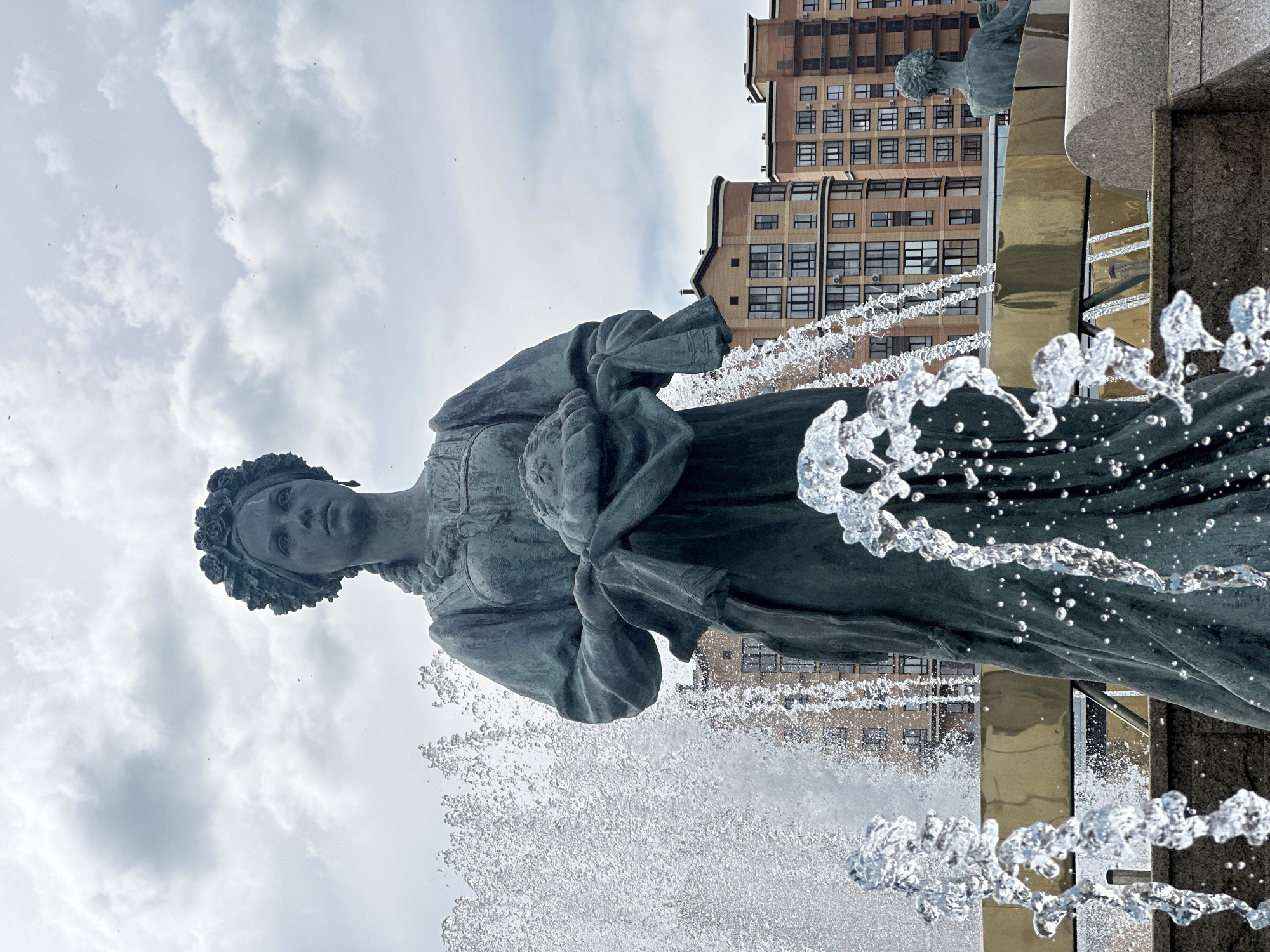 An eye-level, medium shot captures a bronze statue of a woman with a floral headdress and braided hair. She is wearing a traditional dress and holding a bundle of fabric in her arms. The statue is positioned in the foreground, partially obscured by water from a fountain. Behind the statue, a series of multi-story buildings with brick facades and numerous windows are visible under a cloudy sky. Another smaller bronze statue of a seated figure is visible to the right of the main statue. The overall scene is of an outdoor public space, likely a plaza or park, with a fountain and surrounding architecture.