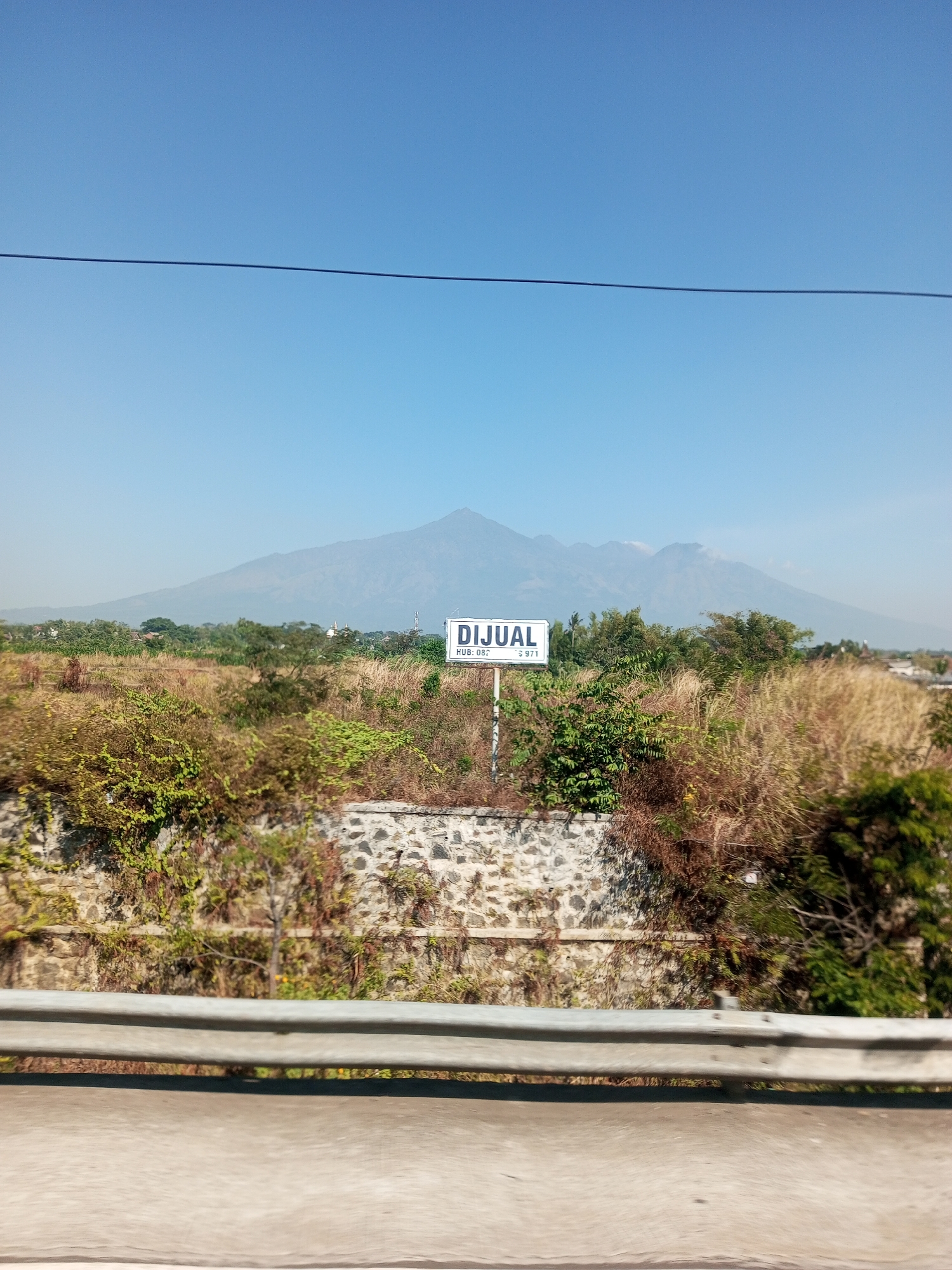 A photograph taken from a vehicle shows a clear blue sky above a landscape featuring a large mountain range in the distance, possibly Mount Semeru in East Java, Indonesia. In the mid-ground, there is a sign that reads "DIJUAL" (Indonesian for "FOR SALE") with "HUB: 082... 971" below it, indicating property or land is available for purchase. The sign is positioned near a low stone wall and surrounded by dry vegetation and scattered trees. In the foreground, a metal guardrail separates the viewer from the road. The overall scene suggests a rural or semi-rural setting with a prominent natural backdrop.