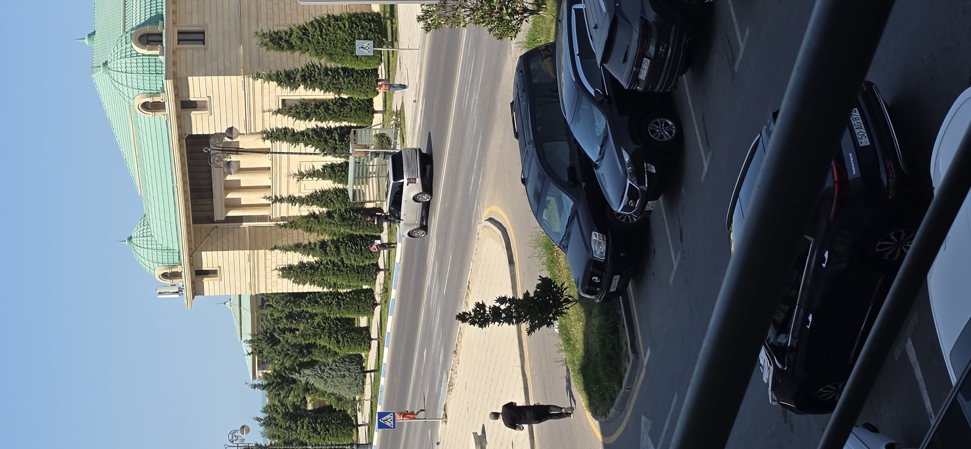 The image depicts a street scene in Sumgayit, Azerbaijan. In the foreground, through the bars of a balcony or railing, multiple dark-colored SUVs are parked. The license plate of one of these SUVs reads "50-AG-874". In the middle ground, a silver SUV is driving on the road. Further back, a sidewalk with a pedestrian crossing is visible, with a few people walking. On the other side of the road, a building with light-colored stone facade and a green dome-shaped roof stands, surrounded by numerous tall, dark green evergreen trees. The sky is clear and blue.