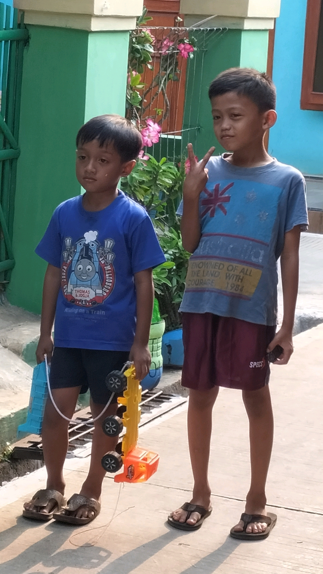 Two young boys stand outdoors on a paved surface. The boy on the left wears a blue t-shirt featuring an image of Thomas the Tank Engine and black shorts. He holds a blue plastic bucket and a yellow toy truck with black wheels. He wears brown flip-flops. The boy on the right wears a blue t-shirt with a graphic design and maroon shorts. He makes a peace sign with his right hand and wears black flip-flops. The background includes a teal-colored wall, a green metal fence, and foliage with pink flowers. The scene appears to be in Subah, Indonesia.