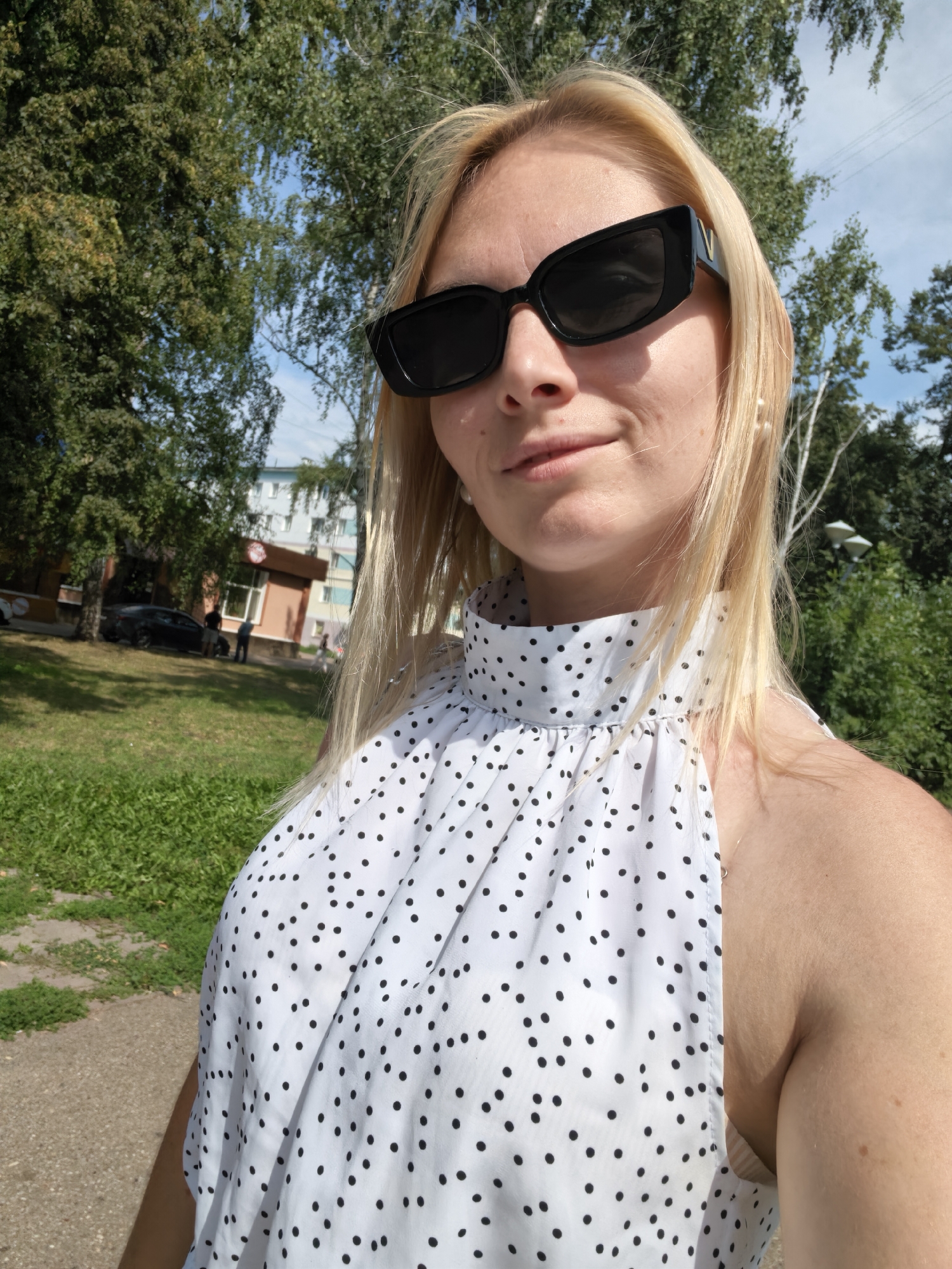 A blonde woman is taking a selfie outdoors in Sterlitamak, Russia. She is wearing large, black, cat-eye sunglasses and a white halter top with black polka dots. Her hair is blonde and falls past her shoulders. She is smiling slightly and looking towards the camera. Behind her, there is a green grassy area with some trees and a sidewalk. In the background, there are buildings and a parked car. The sky is blue with some white clouds. The lighting suggests it is a sunny day.