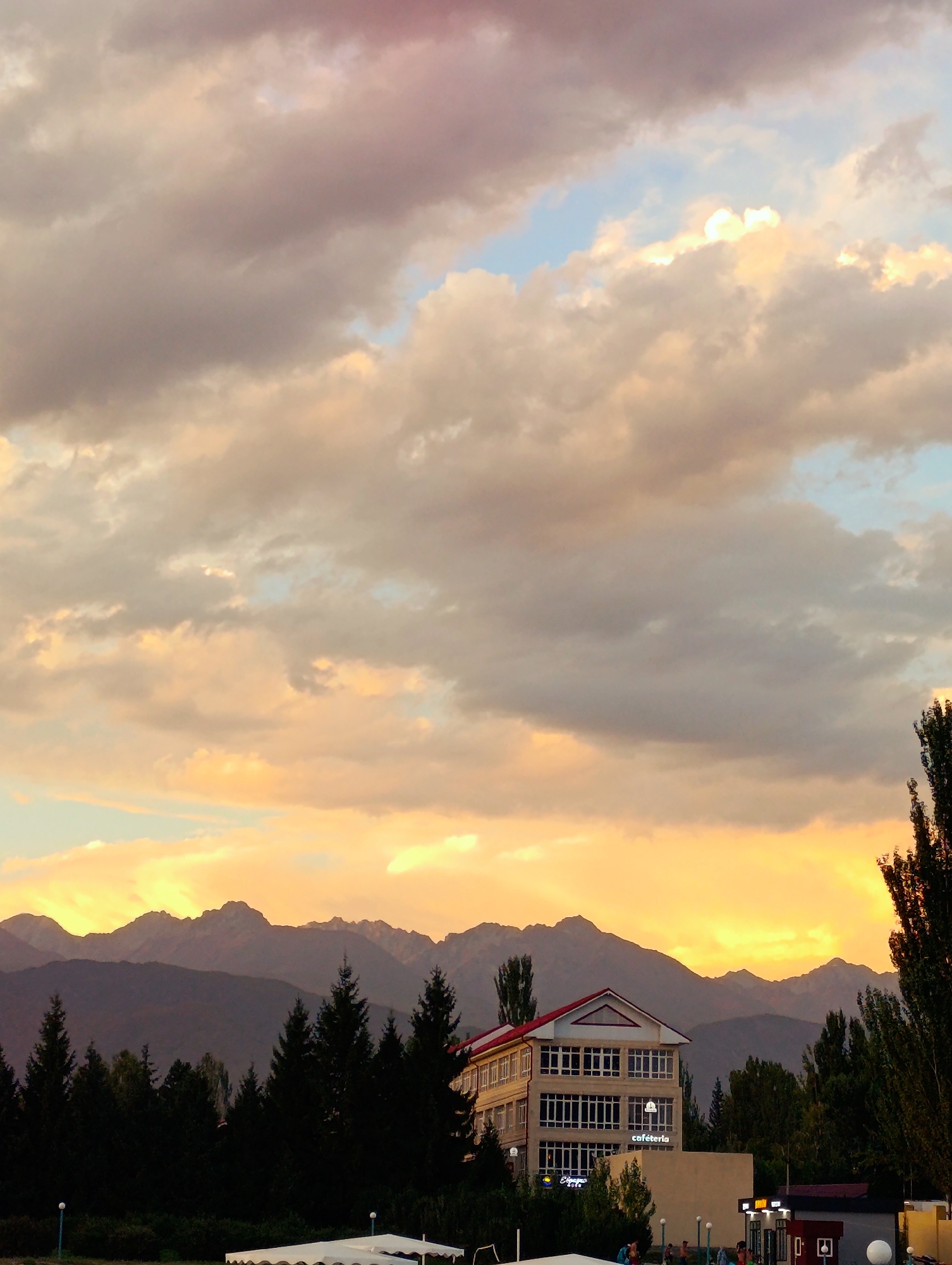 This is a vertical, eye-level, medium shot photograph of a building and mountains at sunset in Bayet, Kyrgyzstan. The sky is filled with dramatic clouds that are illuminated by the setting sun, creating a warm orange and yellow glow. In the foreground, a multi-story building with a red roof stands prominently, its windows reflecting the colors of the sky. The building is surrounded by dark green trees, and beyond them, a range of majestic mountains stretches across the horizon. The overall scene conveys a sense of natural beauty and tranquility.